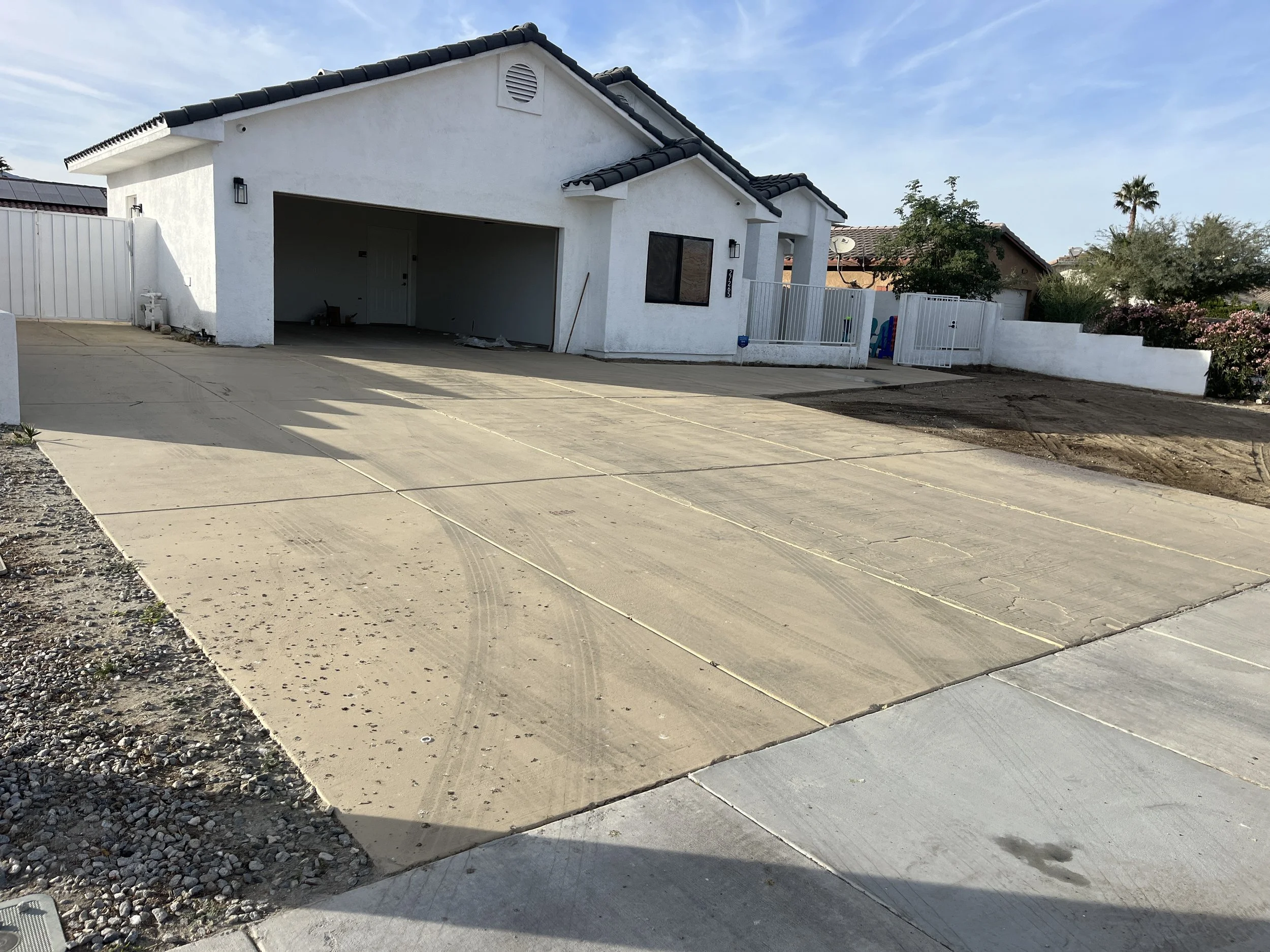 Newly paved driveway leading to a white house with a garage, surrounded by a white fence and desert landscaping, with neighboring houses and desert plants in the background.