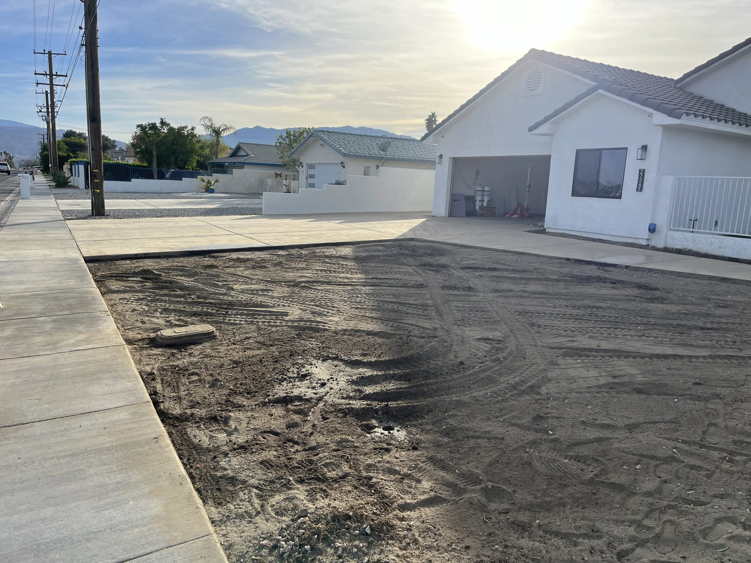 Sidewalk and driveway construction in a residential neighborhood with white houses, utility poles, and mountain views in the background.