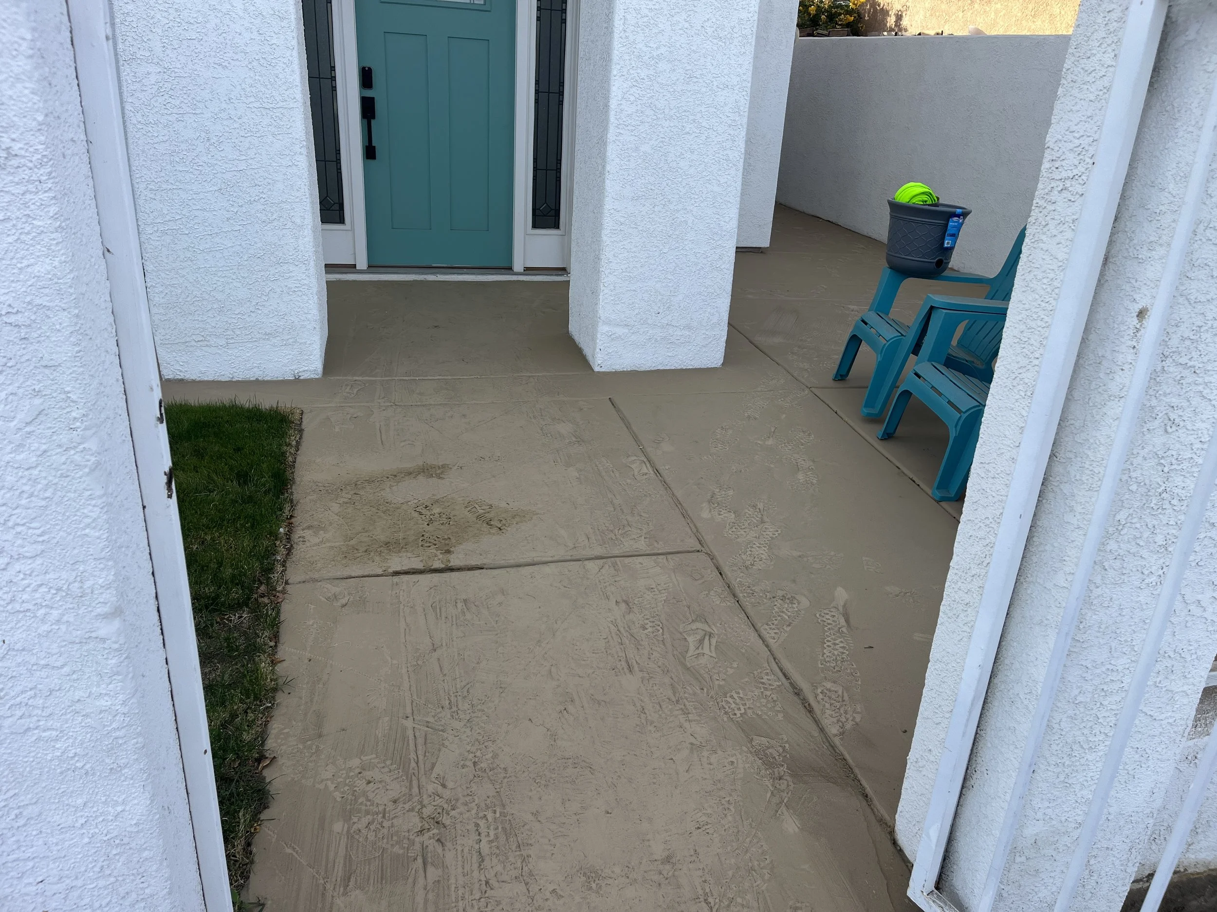 Front porch with a turquoise door, white stucco walls, concrete walkway, two blue chairs, and a black bucket with a green item inside.