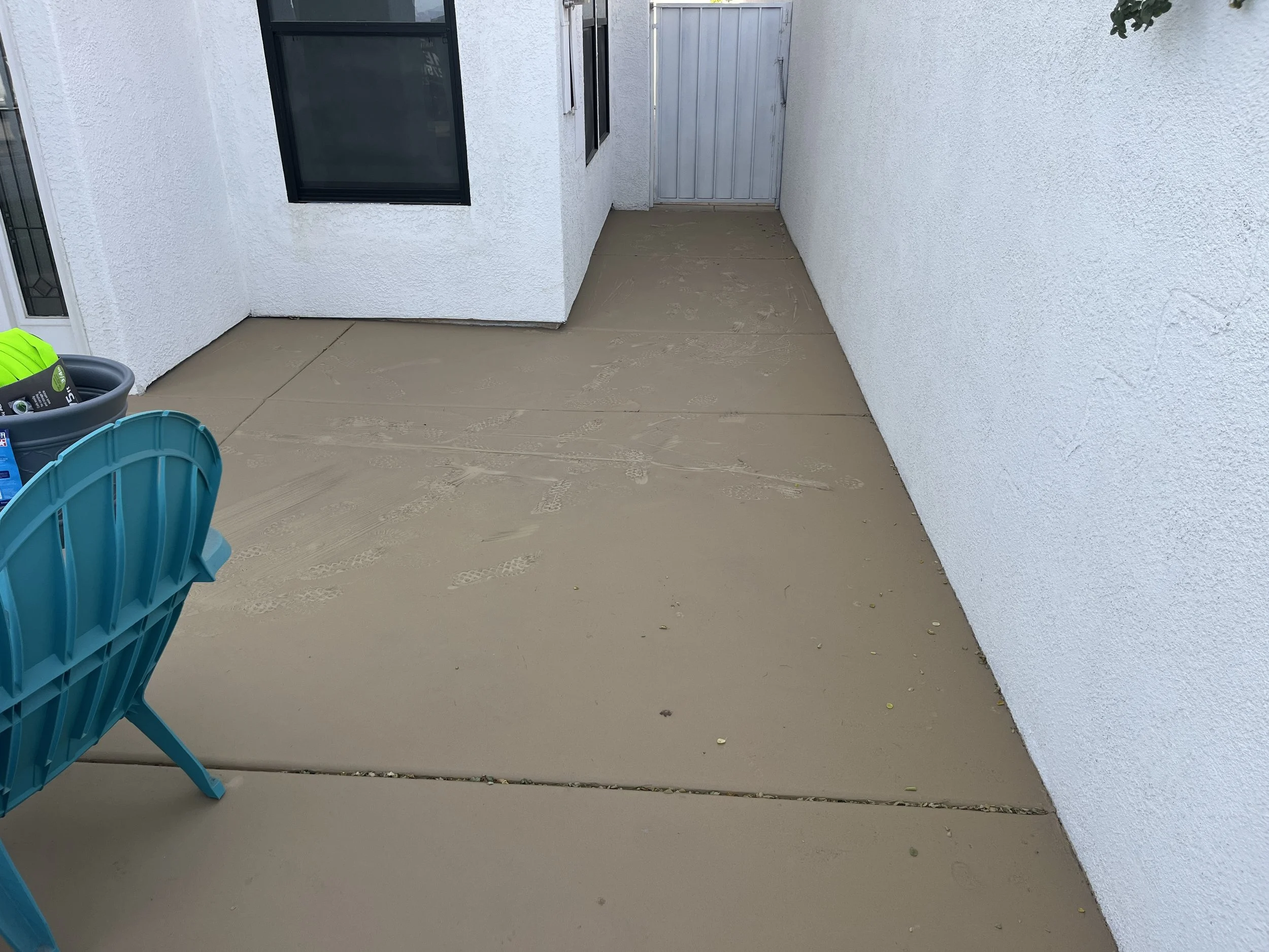 Empty beige concrete patio with white stucco walls, black window frames, and a closed gray metal door. A blue chair and some gardening supplies are visible on the left side.