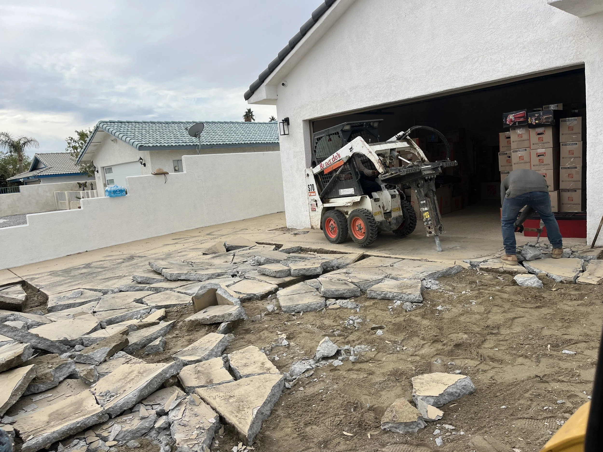 Construction worker and a small skid-steer loader breaking up a concrete patio in front of a garage. Piles of broken concrete are scattered on the dirt ground.