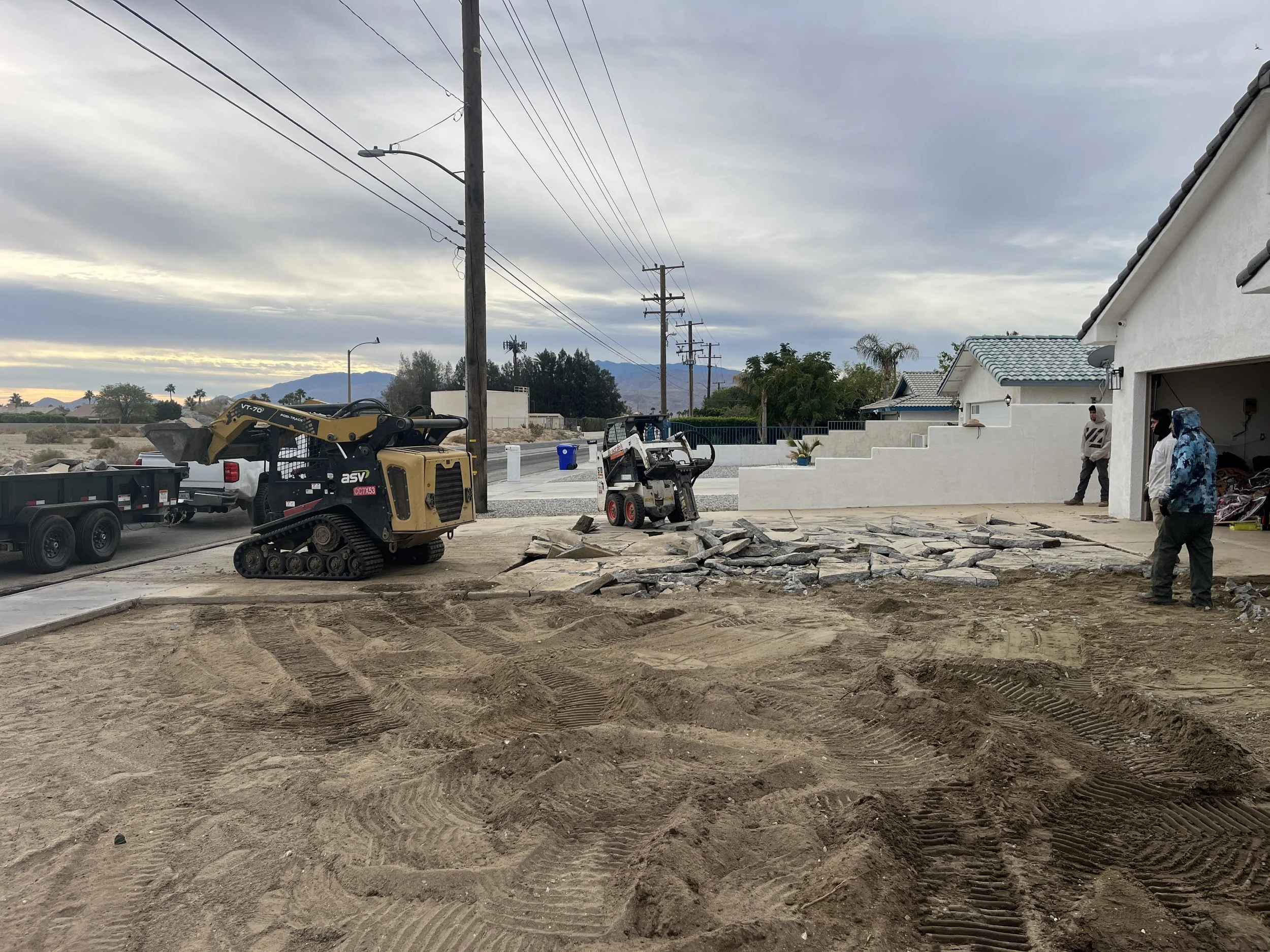 Construction workers and machinery laying stone pavers in the driveway of a white house, with utility poles and a residential neighborhood in the background during cloudy weather.