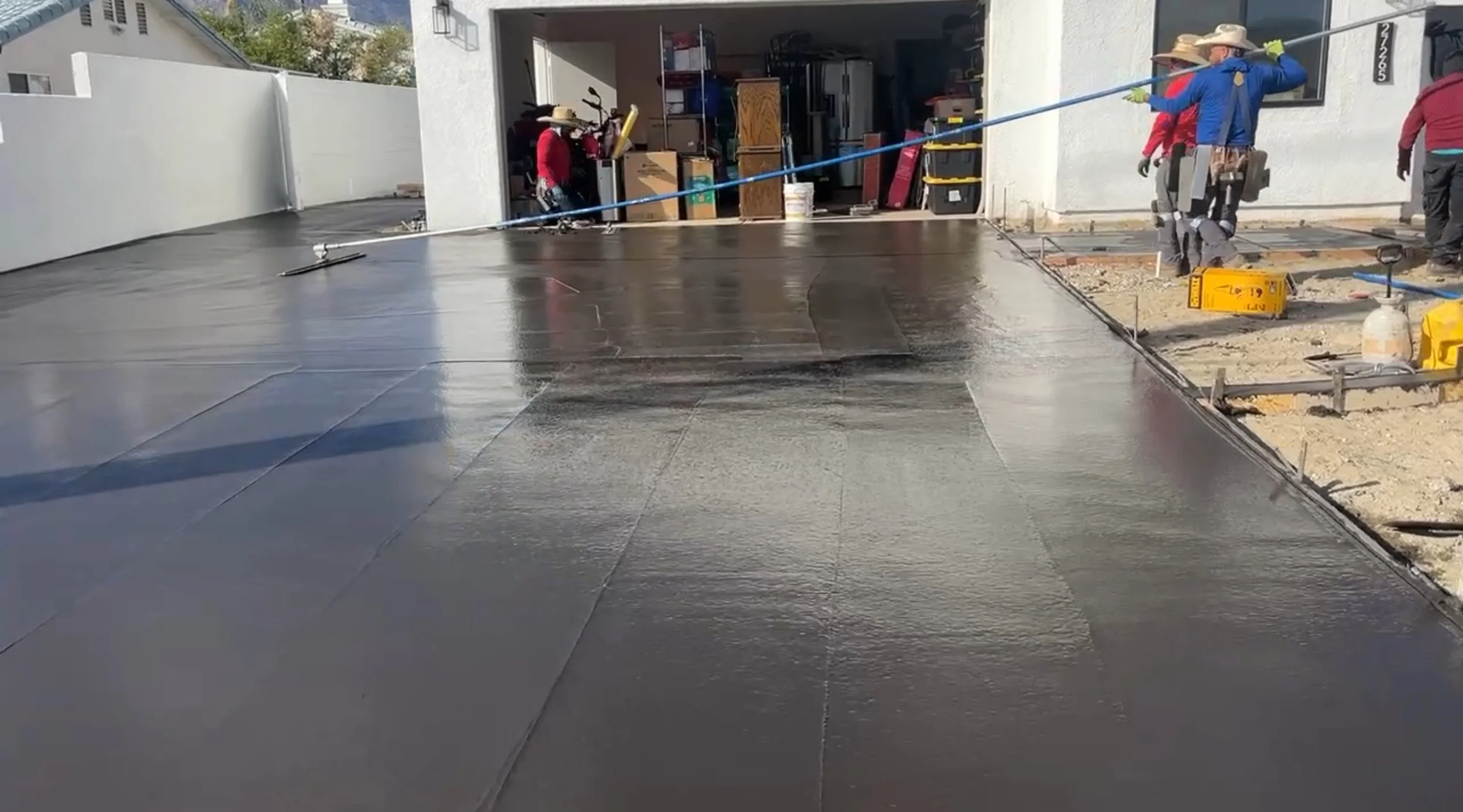 Construction workers applying a fresh coat of black waterproofing to a concrete driveway outside a house.