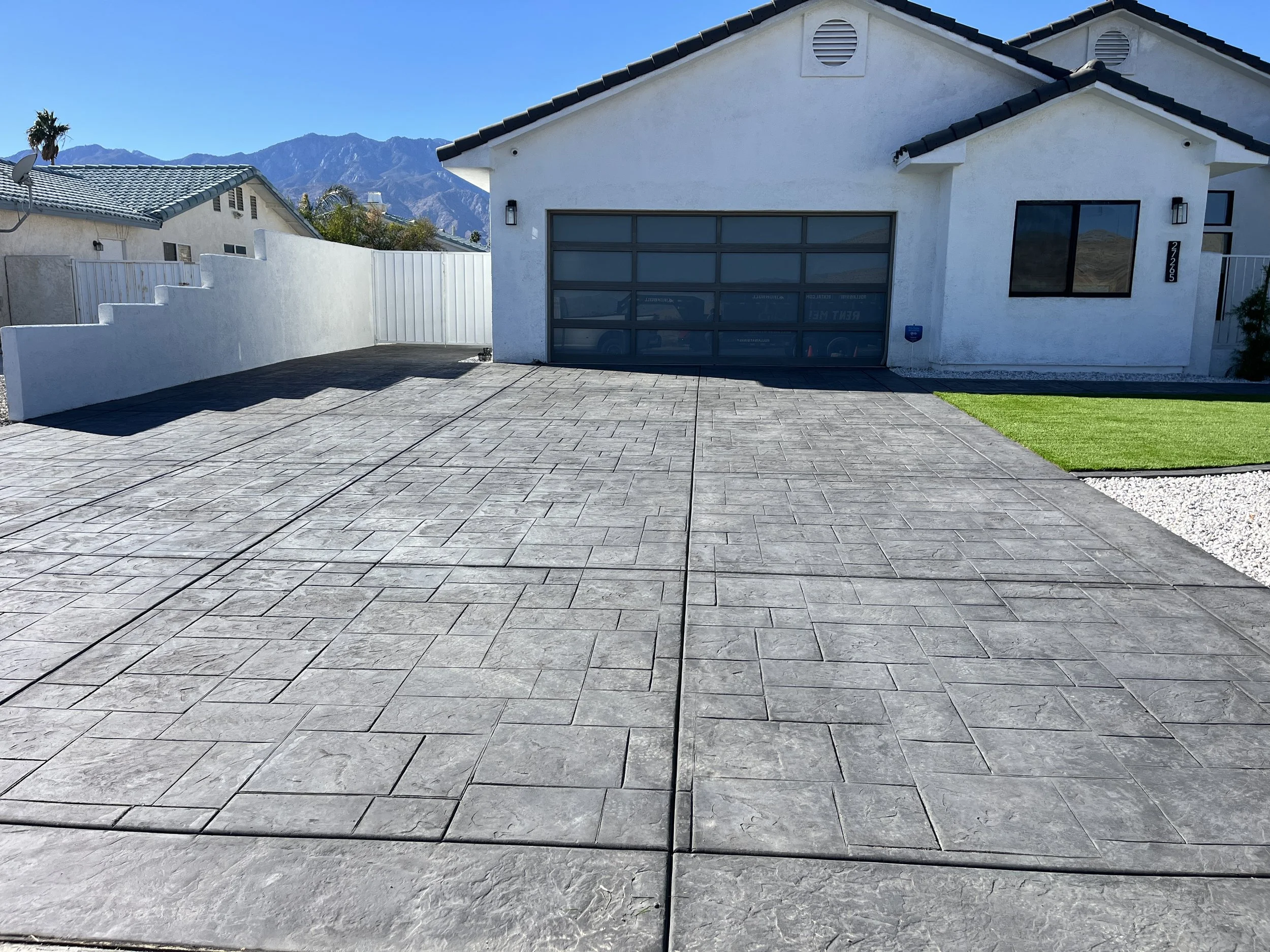 Front view of a white house with a concrete driveway, garage door, and a small patch of green grass on the right, with mountains in the background under a clear blue sky.