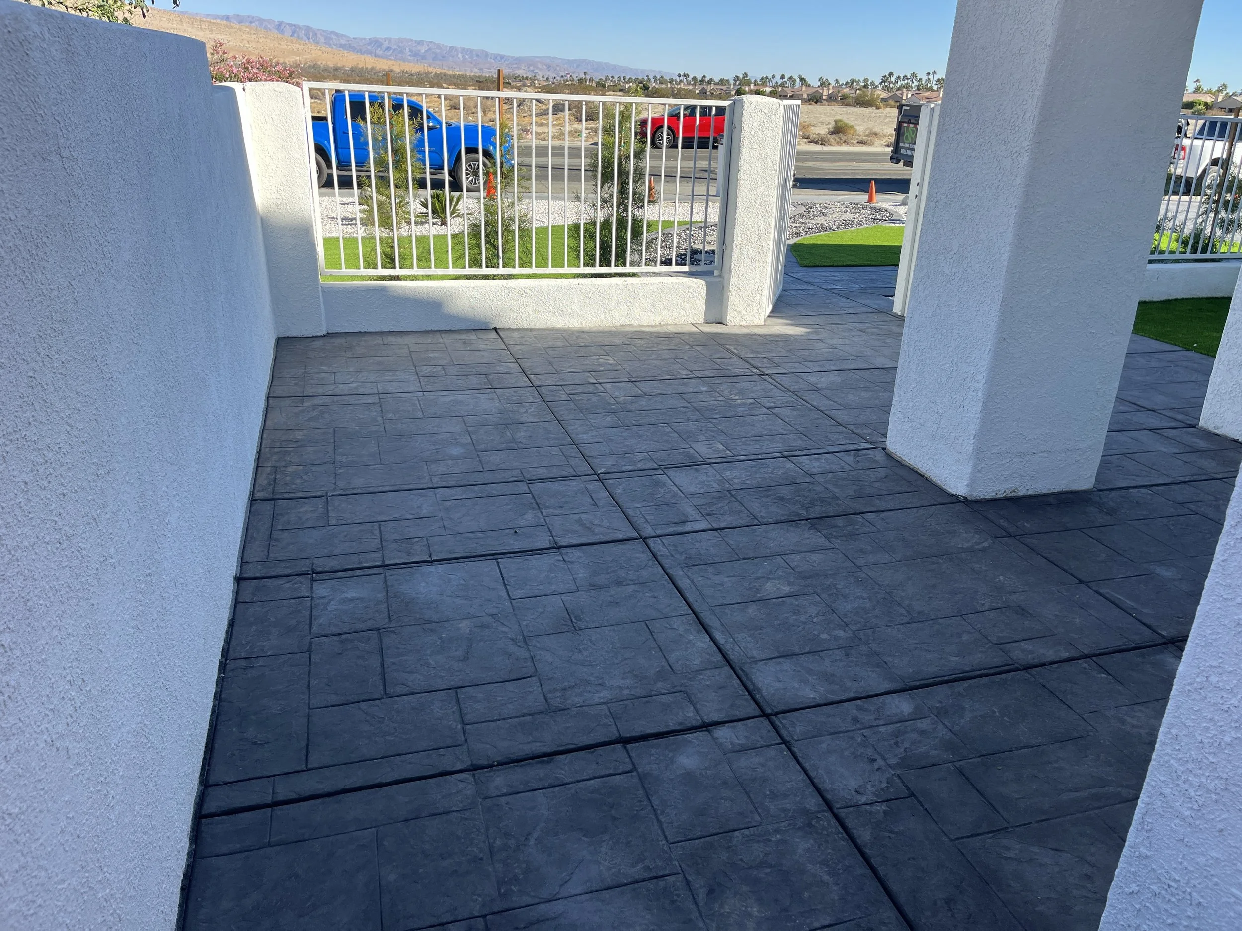 View of a patio with black stamped concrete flooring, white stucco walls, a white metal gate, artificial green grass on the sides, and parking lot with cars and mountains in the background.