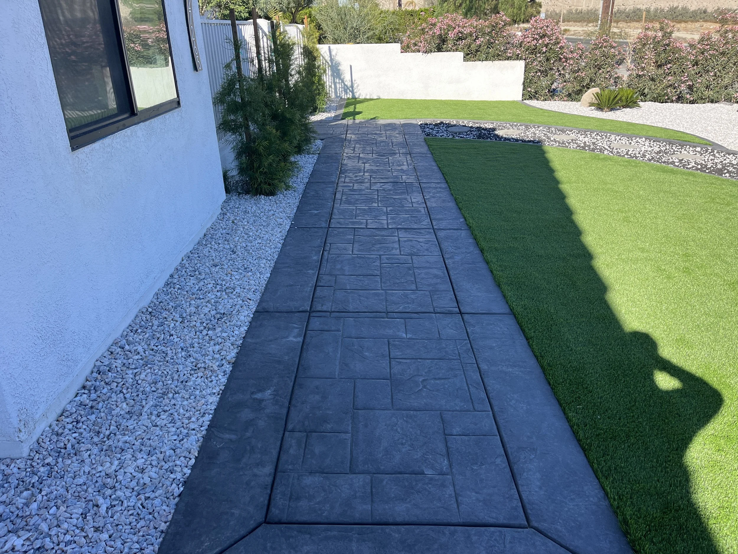 A concrete pathway runs alongside a white stucco house with a window, bordered by white gravel, with green shrubs on one side and a well-manicured lawn with a landscaped area with rocks and bushes in the background.