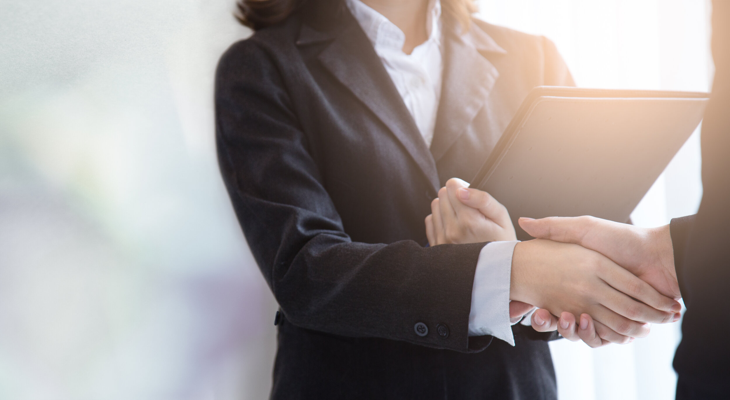 Two people shaking hands, one in a business suit holding a clipboard, inside a bright office.