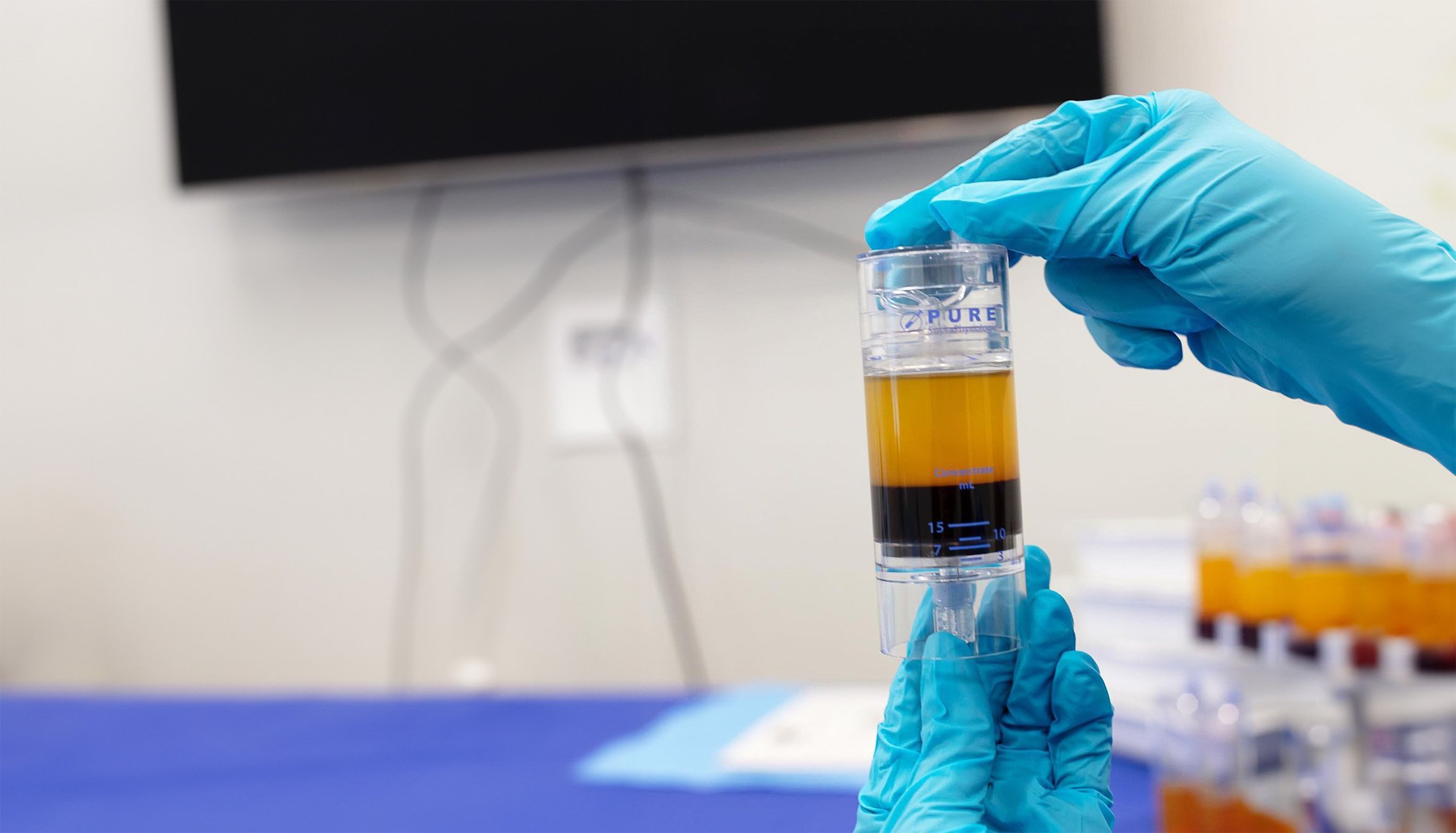 A gloved hand holding a graduated cylinder with liquid, in a laboratory setting.