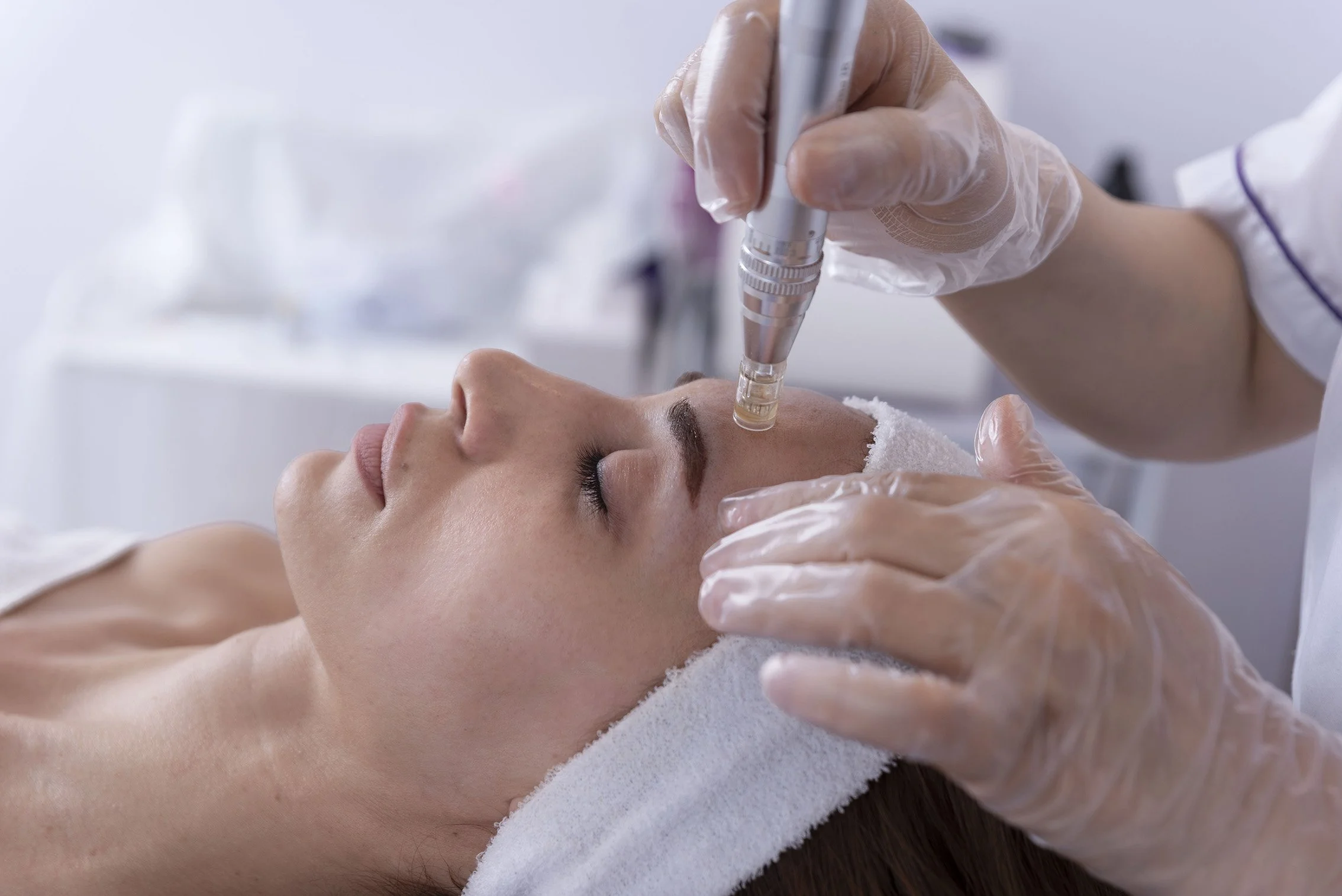 A woman receives a facial treatment with a skincare device on her face, lying down with eyes closed, in a clinical or spa setting.