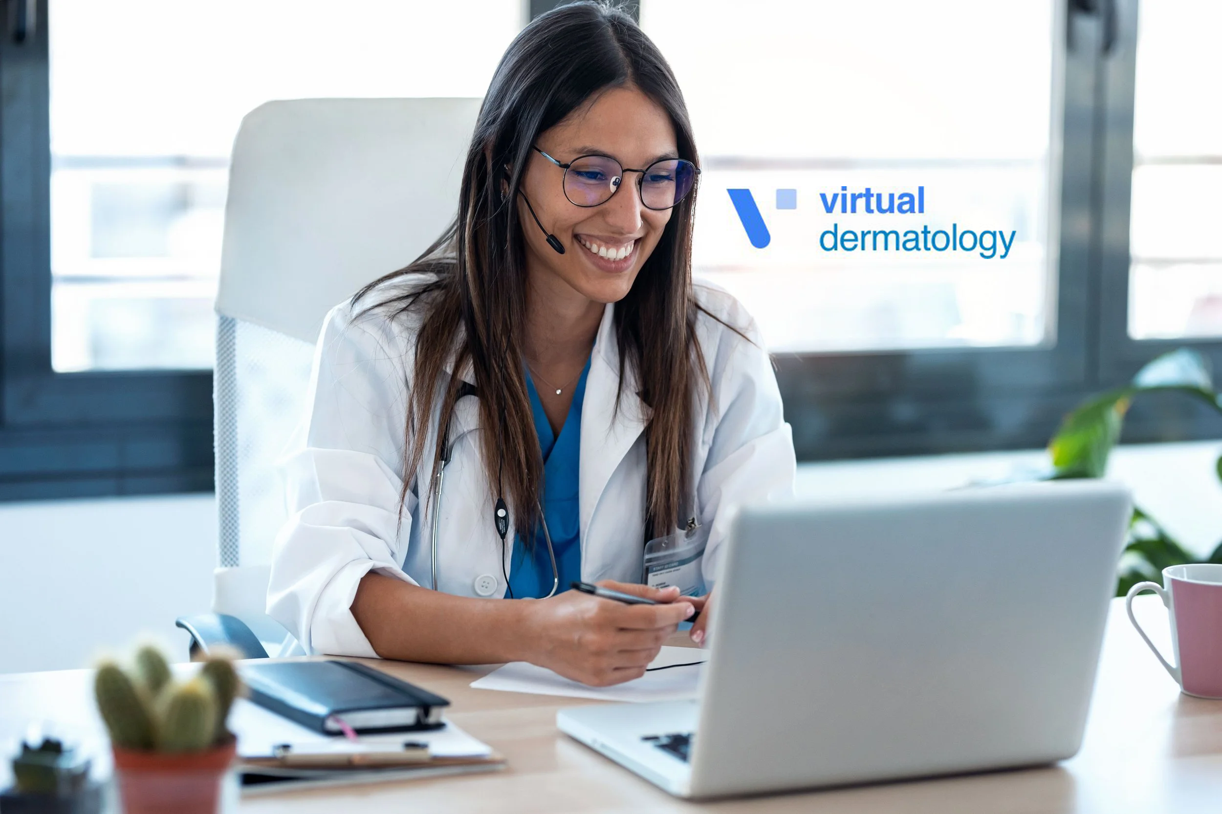 A female doctor with long dark hair, glasses, and a headset is smiling while looking at her phone in an office setting. She is wearing a white coat, and there is a laptop, notebook, and potted plants on the desk. Glass windows are visible in the background, and a logo reading 'virtual dermatology' is displayed on the image.
