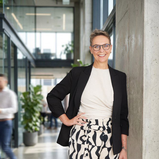 Businesswoman smiling in modern office building, wearing glasses, a black blazer, white top, and patterned pants.