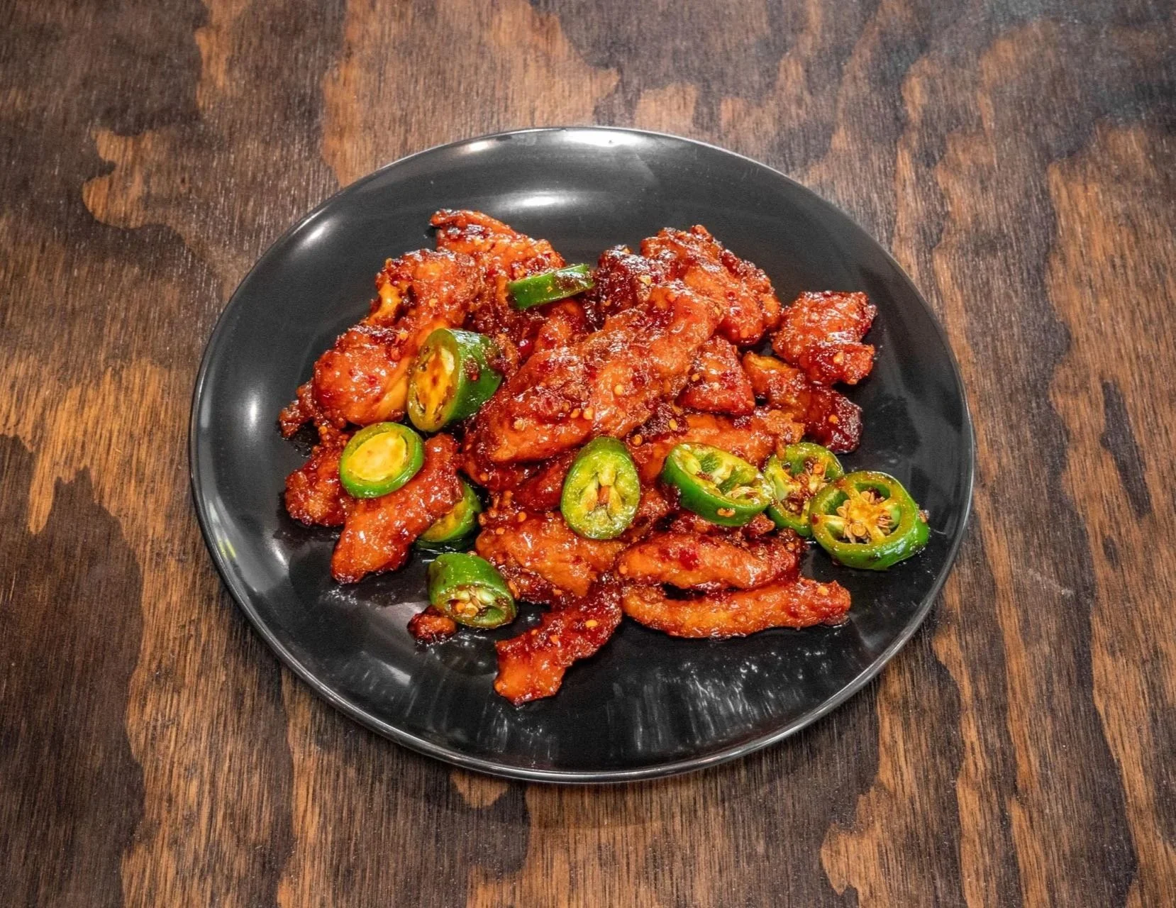 Spicy fried chicken pieces topped with sliced green chili peppers on a black plate, set on a wooden table.
