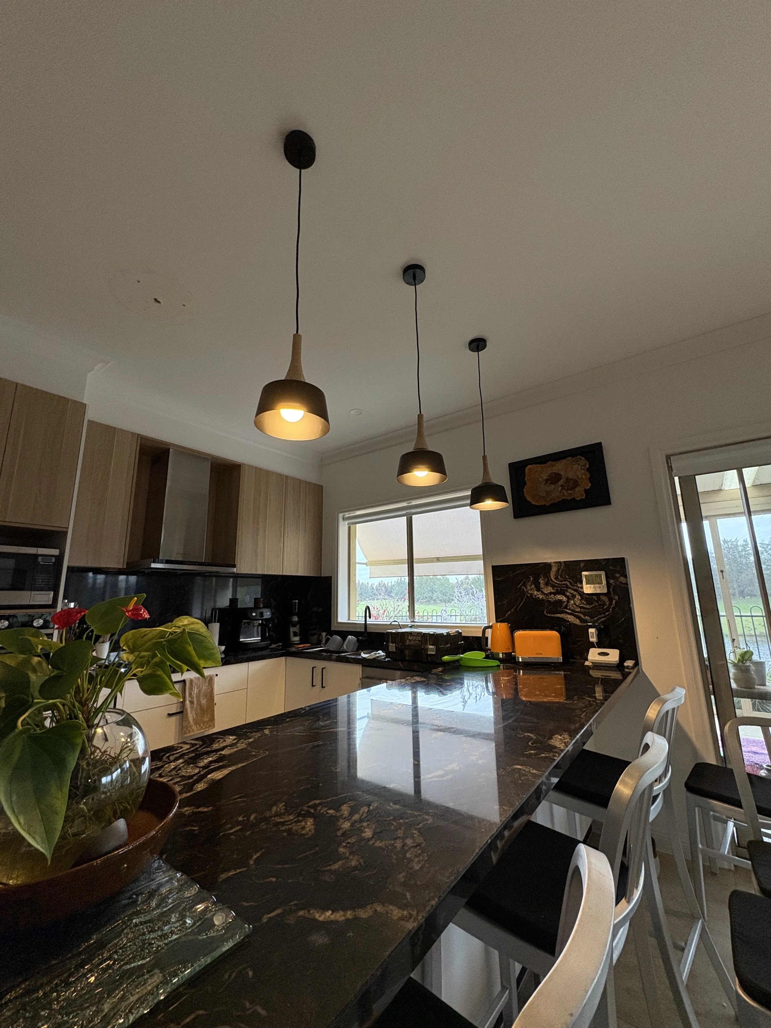 Kitchen with black marble countertop, wooden cabinets, hanging pendant lights, window showing an outdoor view, plants on the counter, and barstools along the counter.