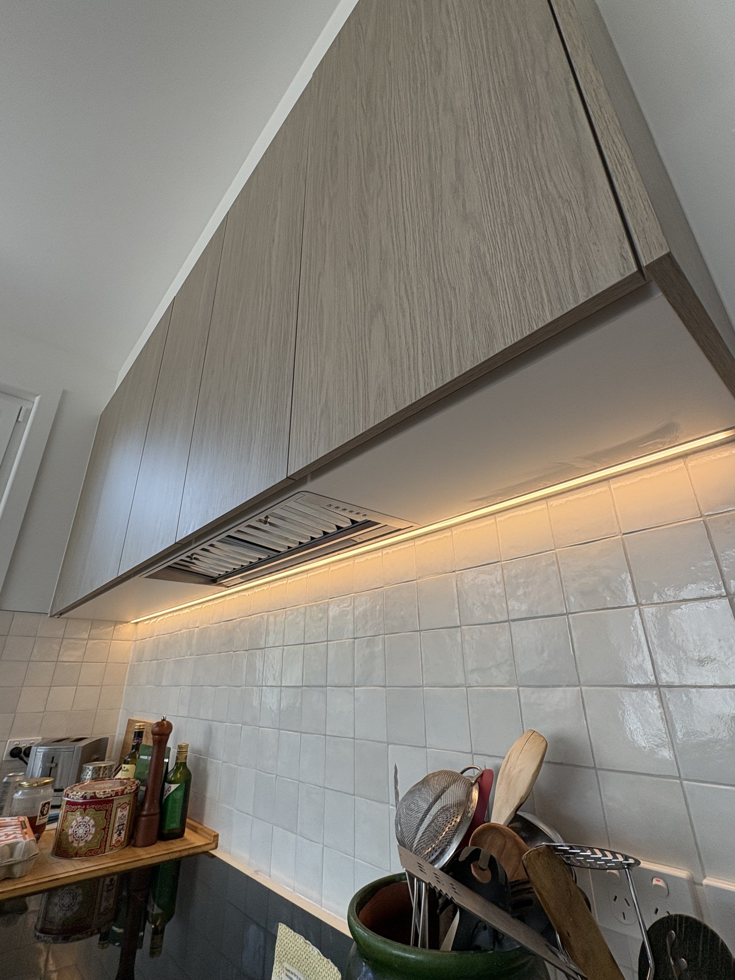 Modern kitchen with wooden cabinet above tiled backsplash, under-cabinet lighting, and various kitchen utensils and bottles on the countertop.