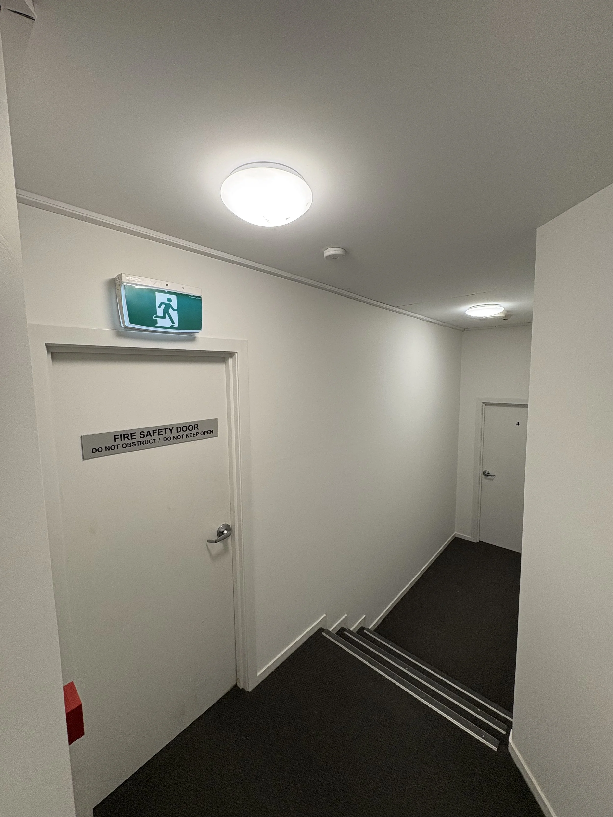 White hallway with two ceiling lights, a fire safety door labeled with a sign, an emergency exit sign above the door, and a staircase leading downward.