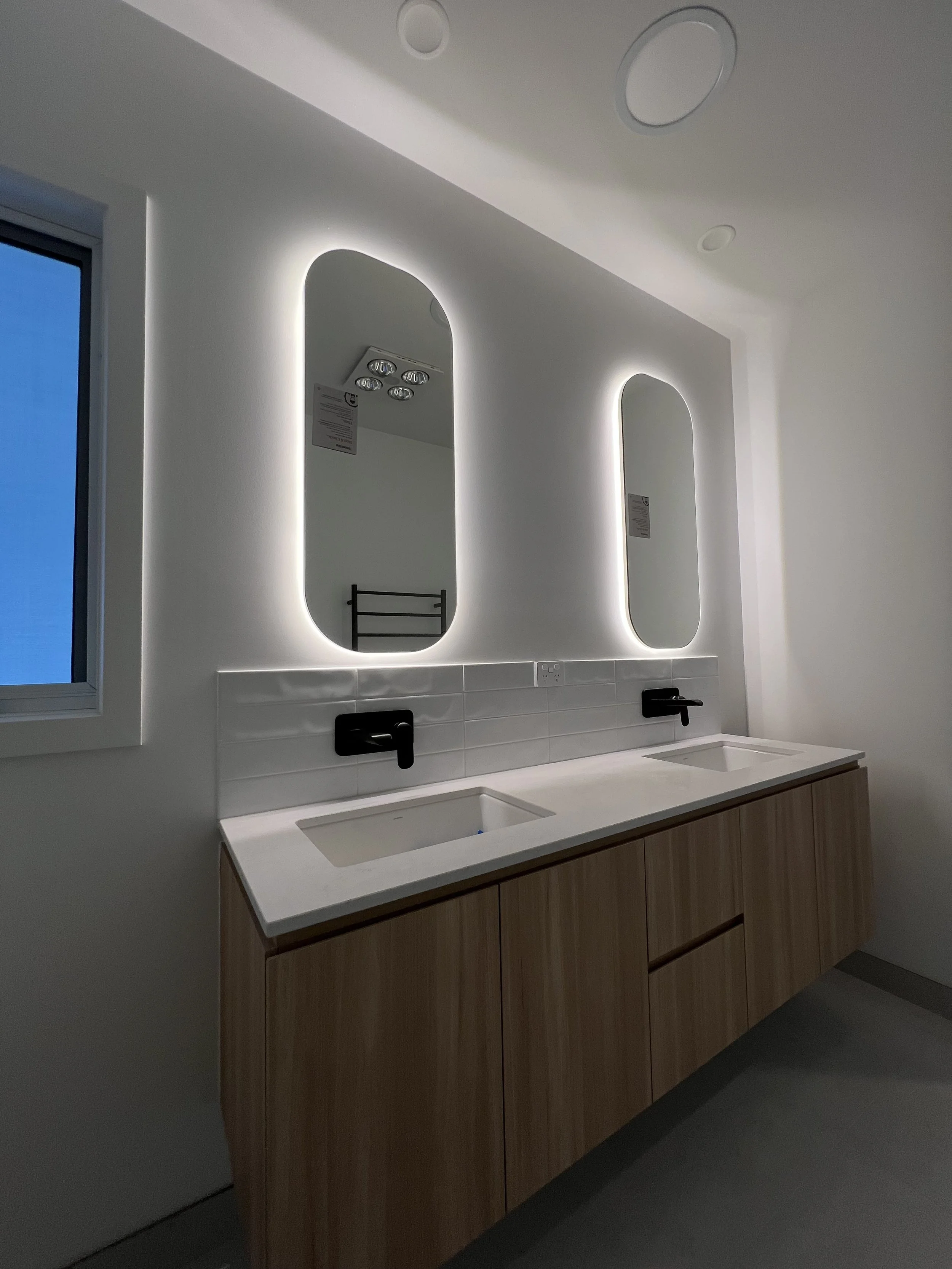 Modern bathroom vanity with dual sinks, black faucets, backlit mirrors, and a wooden cabinet, illuminated with LED lighting.
