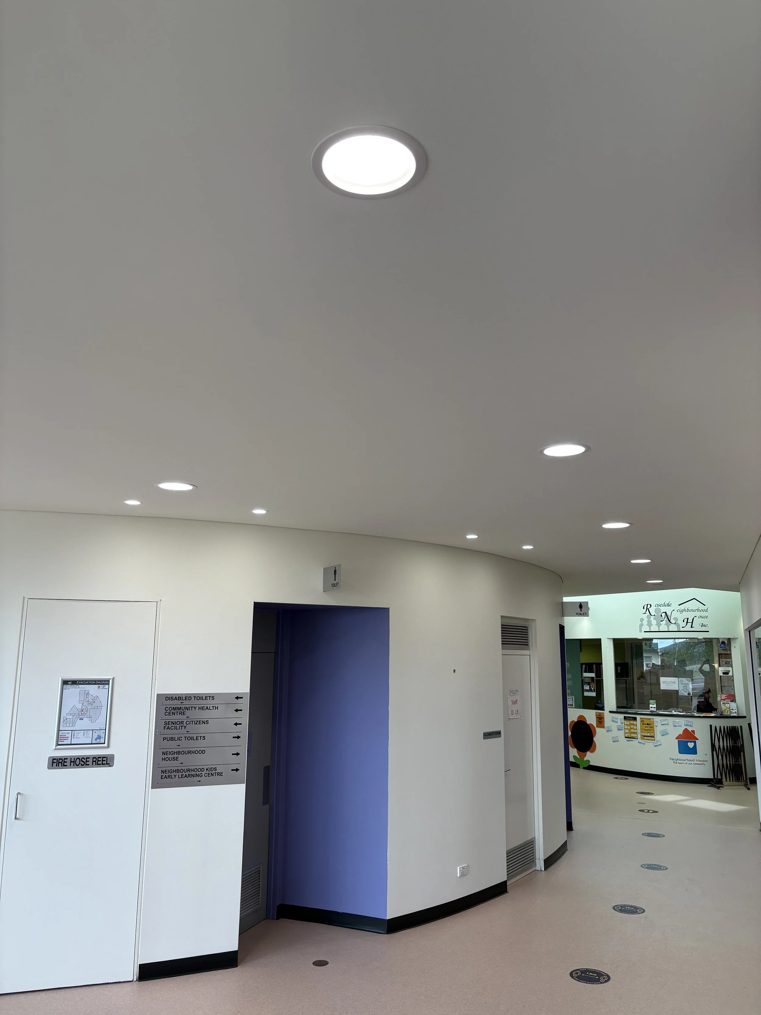 Interior view of a public building with ceiling lights, a door with sign for toilets, and a reception area in the background.