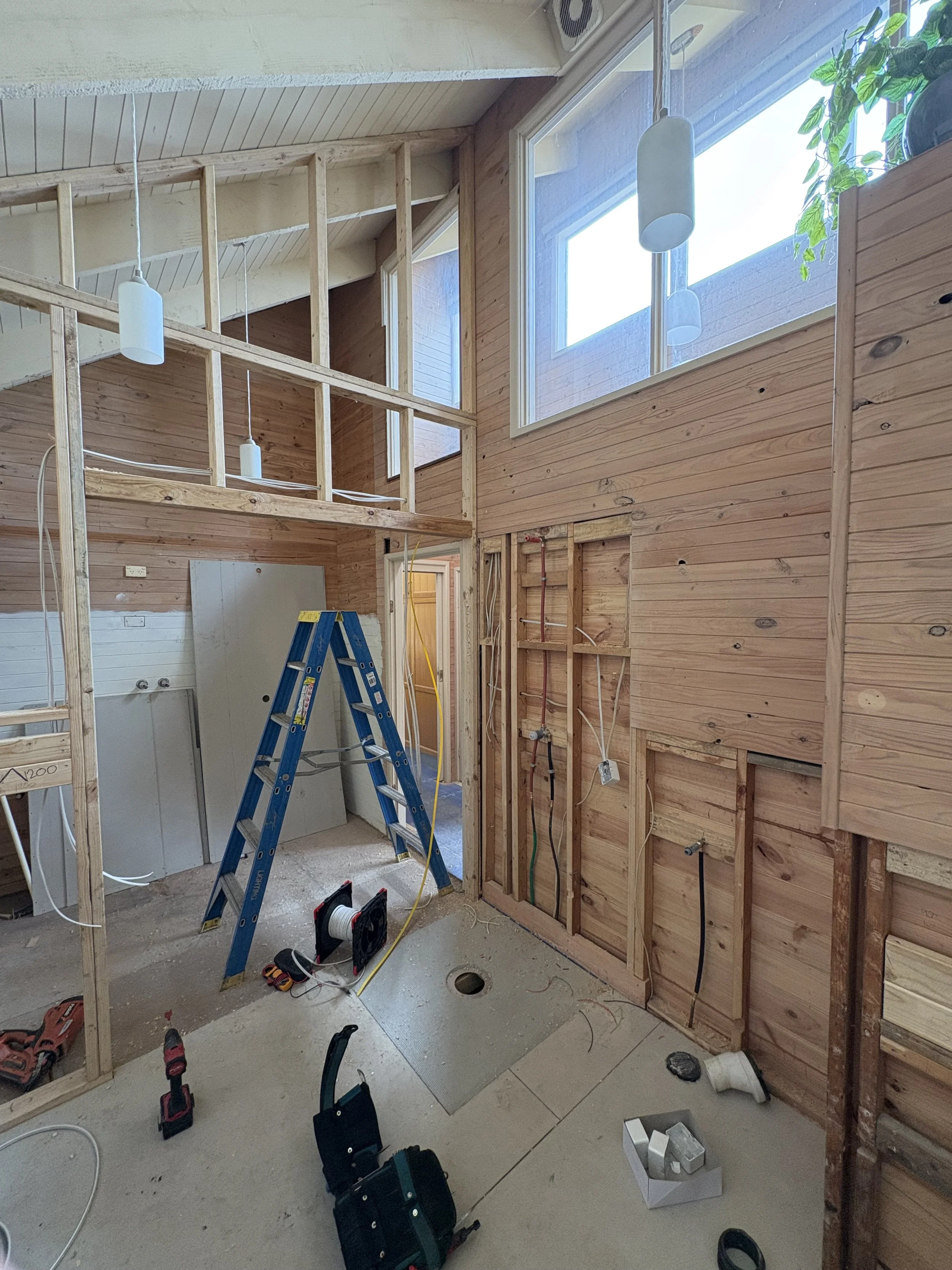 Interior of a house under construction with exposed wooden framing, a blue ladder, construction tools, electrical wiring, and unfinished walls.