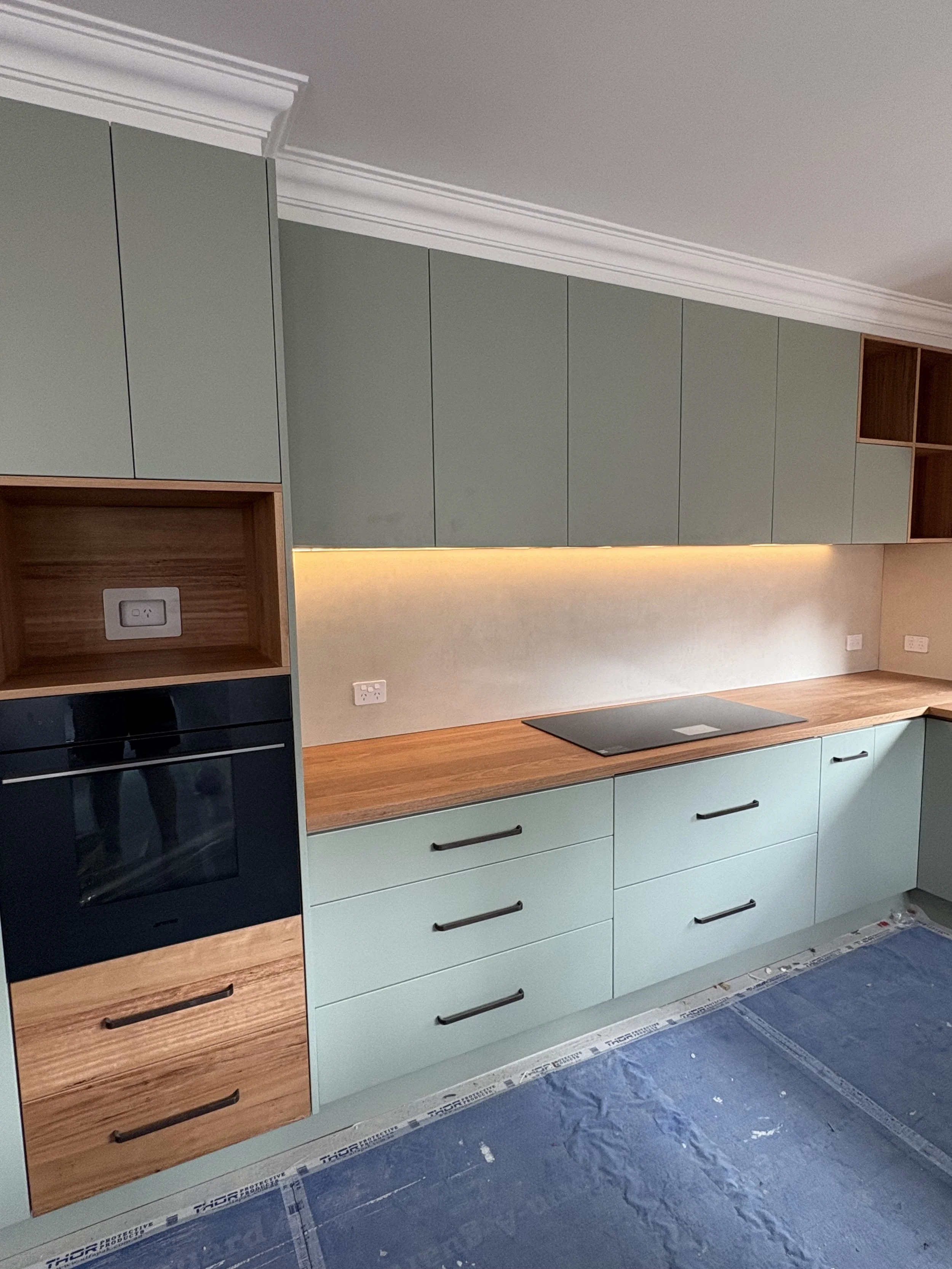 Modern kitchen with green cabinets, wooden countertop, built-in oven, electric cooktop, and open shelving, with electrical outlets and under-cabinet lighting.