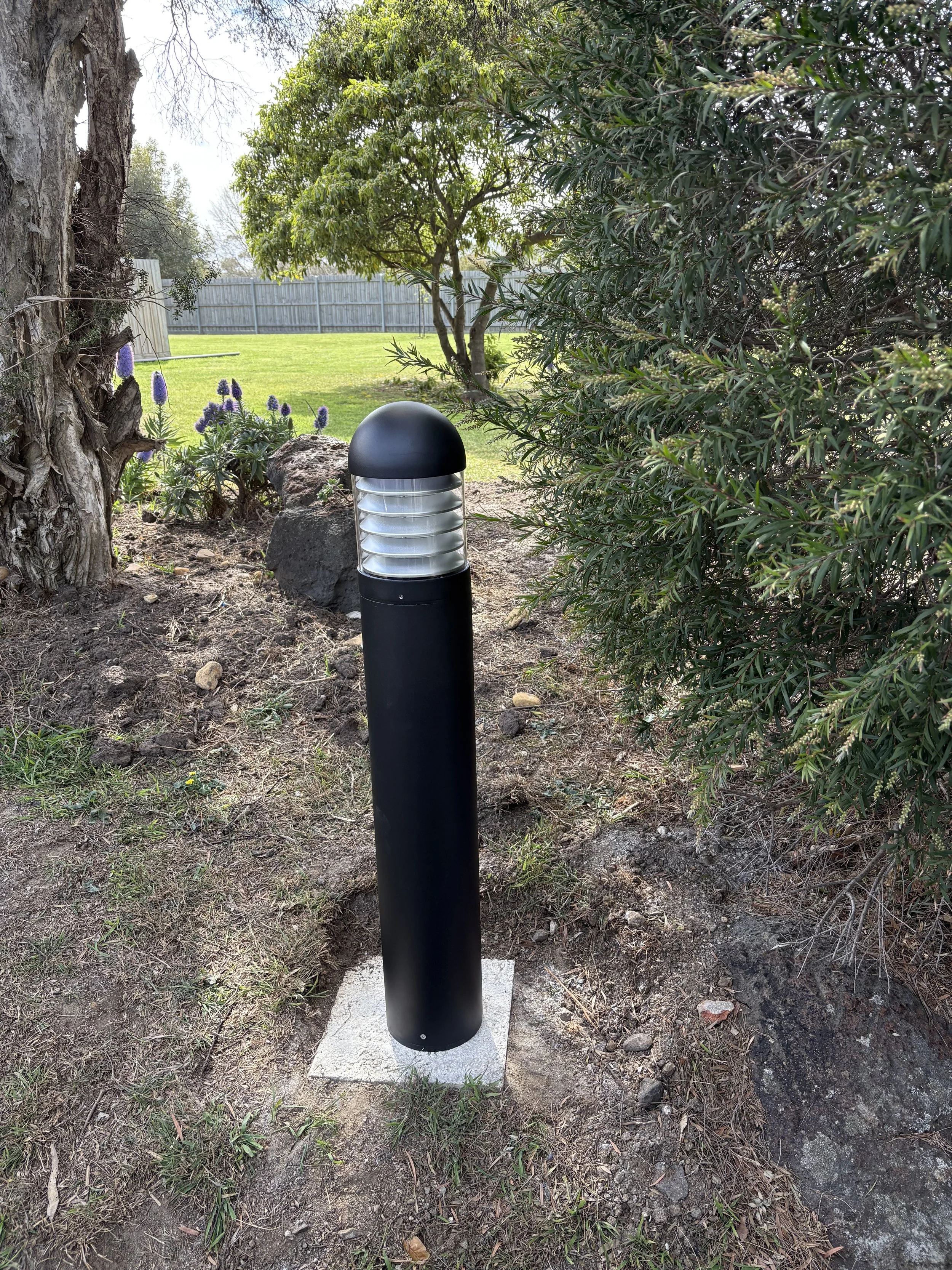 Outdoor garden with a black modern post light fixture on a concrete base, surrounded by dirt and plants, with a fence and trees in the background.