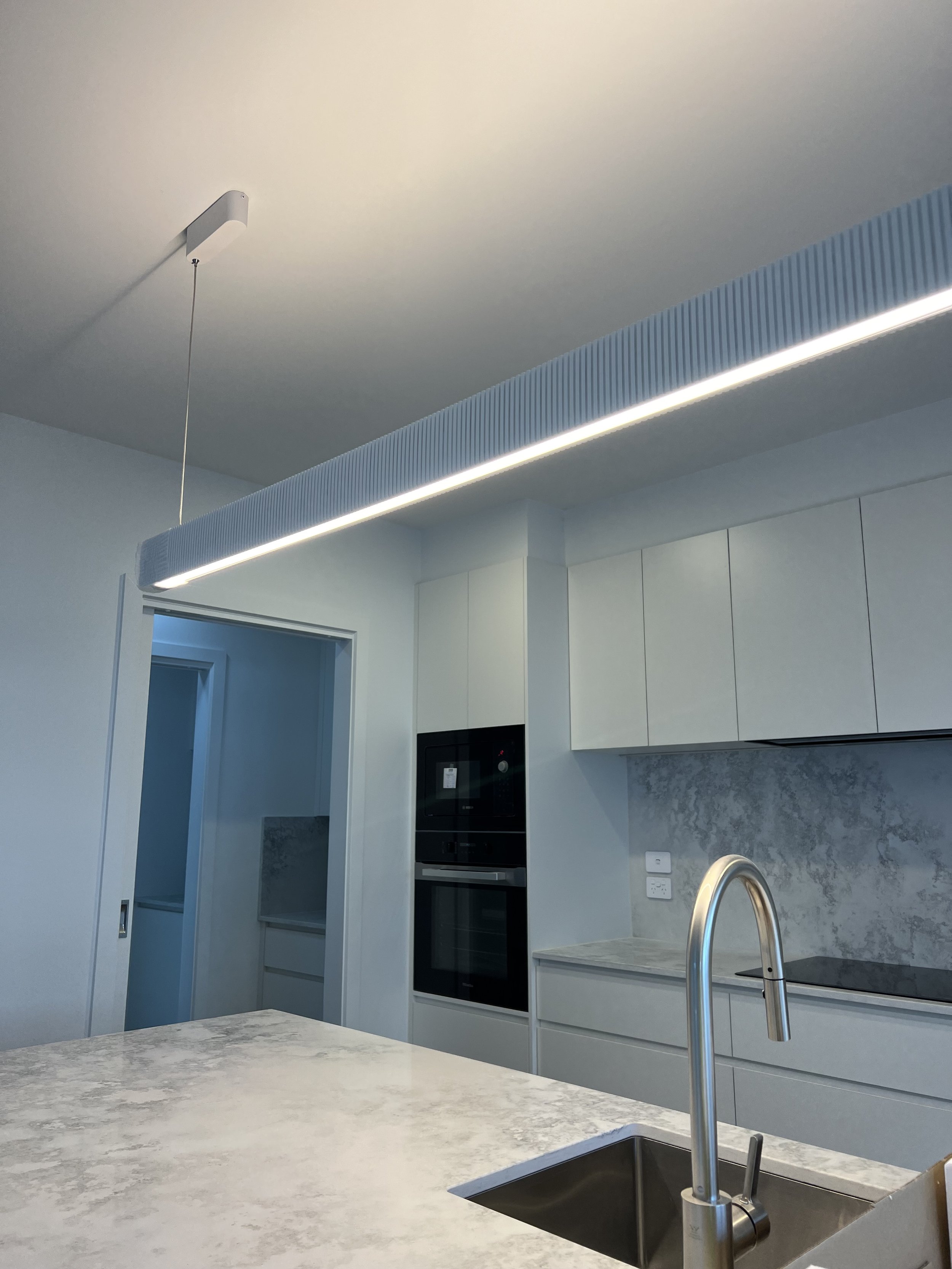 Modern kitchen with a marble countertop island, white cabinets, stainless steel faucet, built-in oven, and minimalist lighting fixture.