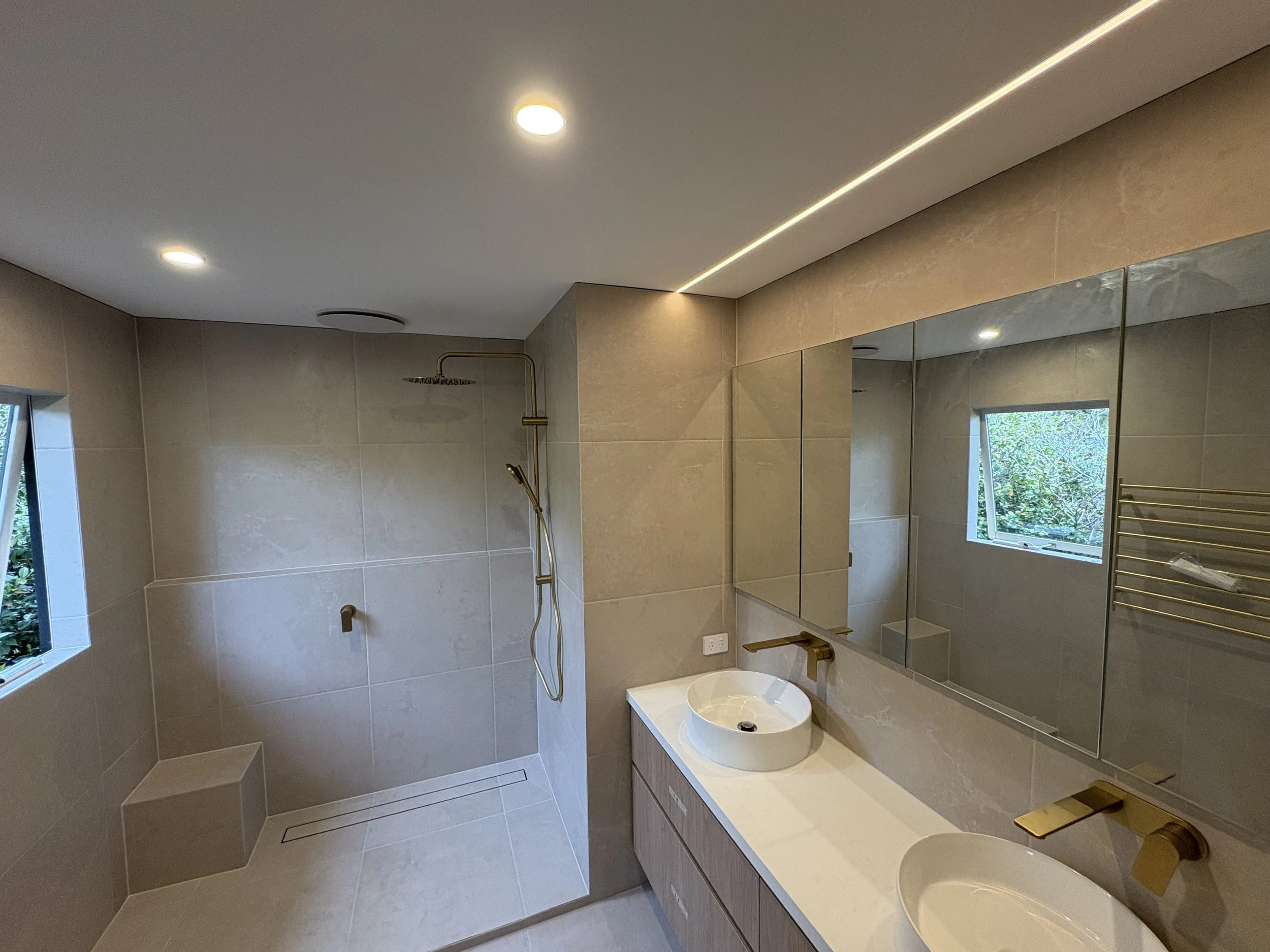 Modern bathroom with beige tiled walls and floor, walk-in shower with rainfall and handheld showerheads, double vanity with two vessel sinks, large mirror, window showing greenery outside, gold fixtures, and built-in lighting.