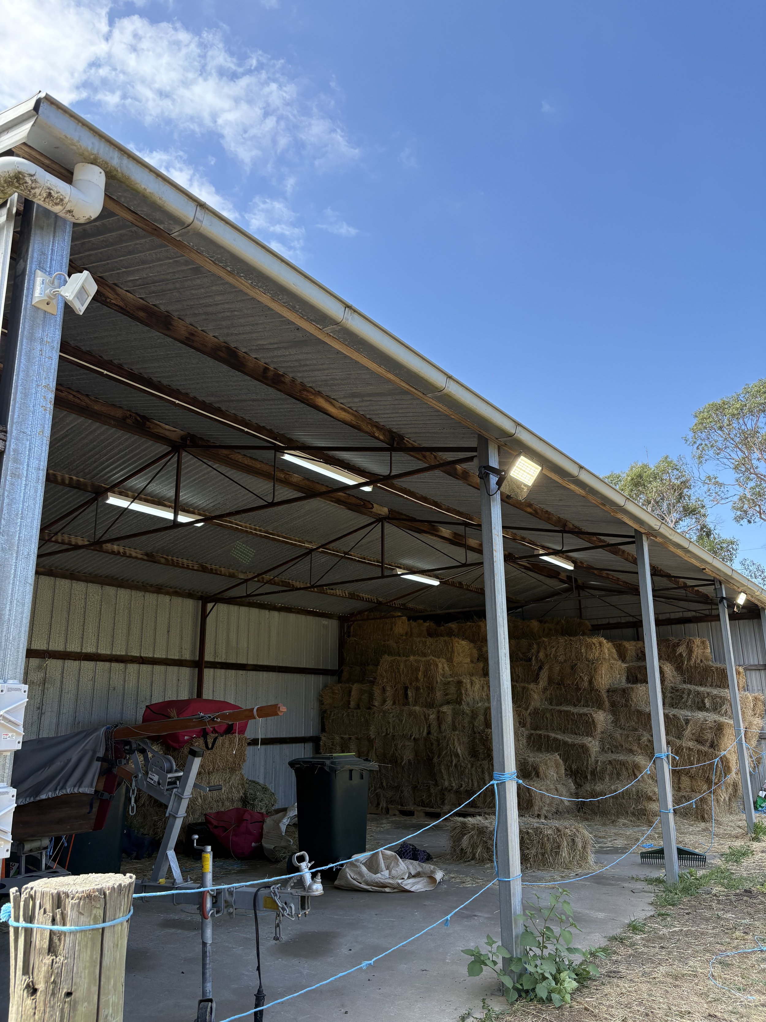Barn with a metal roof and walls, filled with hay bales stacked inside. Equipment and supplies are scattered in front, with a blue rope barrier, and z1 trees visible in the background under a partly cloudy sky.
