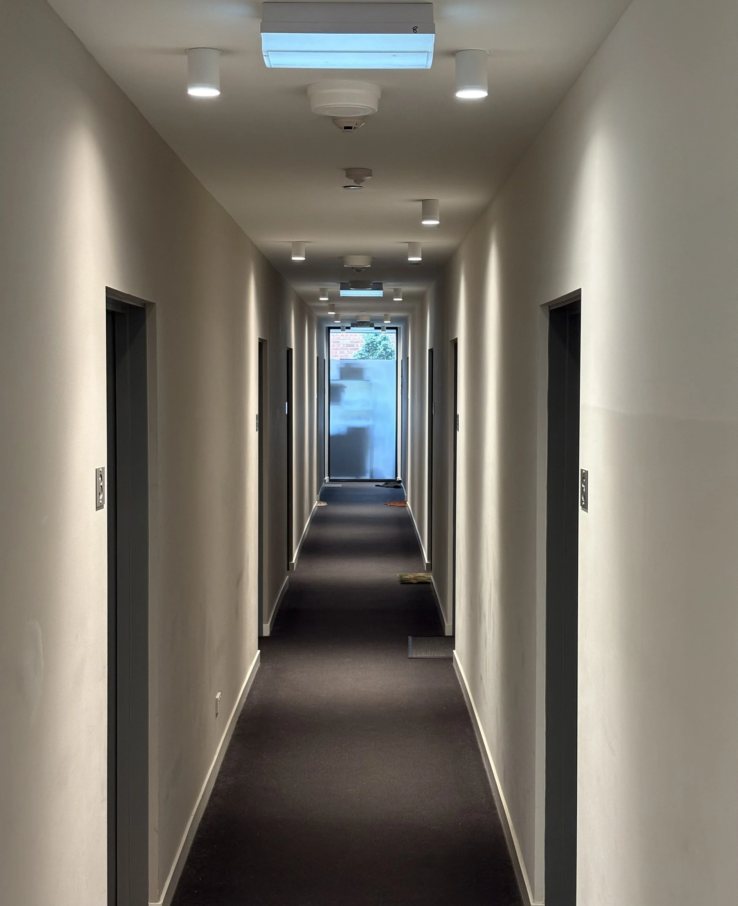 Empty hotel hallway with closed doors on either side, ceiling lights, and a glass door at the end, with some doormats on the dark carpet.
