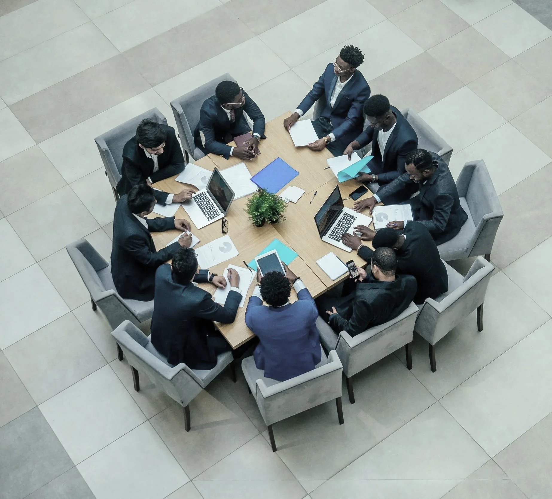 A top-down view of twelve professionals in business suits gathered around a rectangular conference table with laptops, tablets, documents, and a small potted plant, engaged in a meeting in a modern office setting.