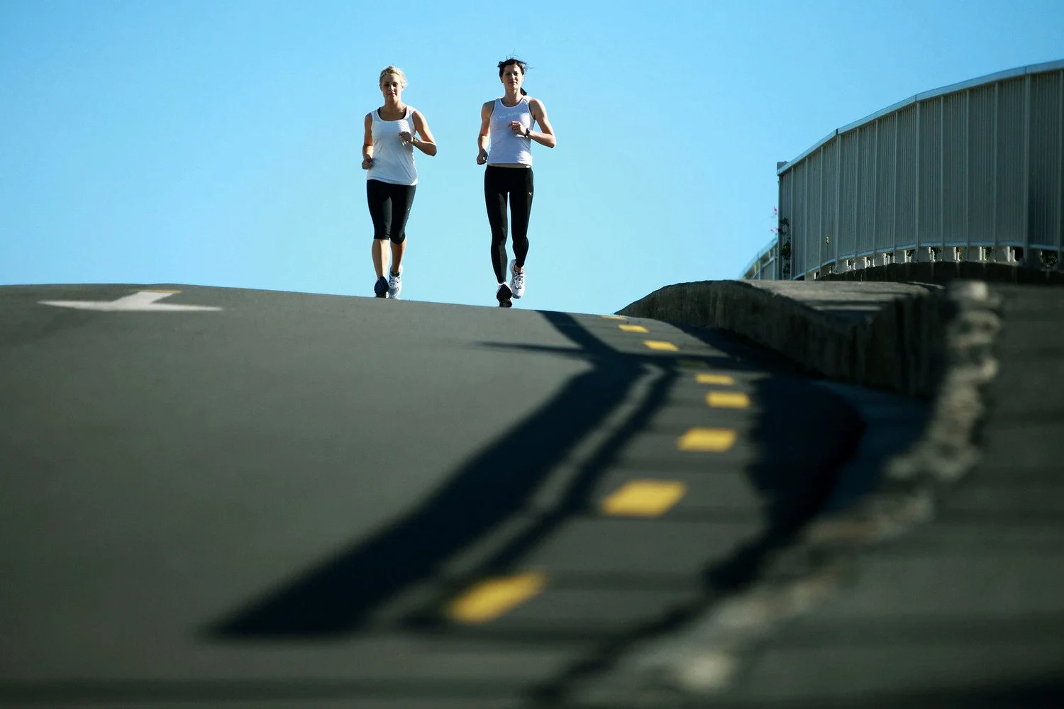 Two women jogging on a slightly uphill road with a blue sky overhead and a metal fence on the right side.