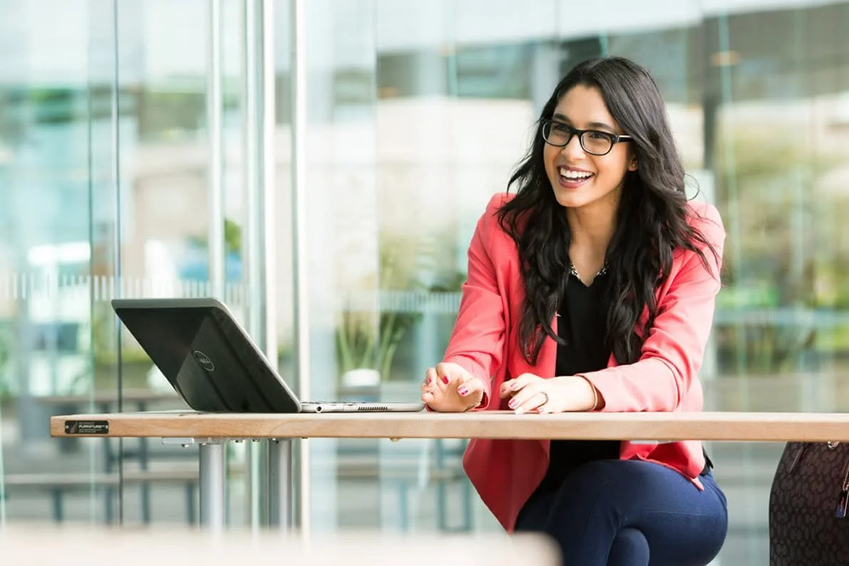 A woman with long dark hair, glasses, and a pink blazer sitting at a table with a laptop, smiling and engaging in a conversation or video call in a bright, modern office environment.