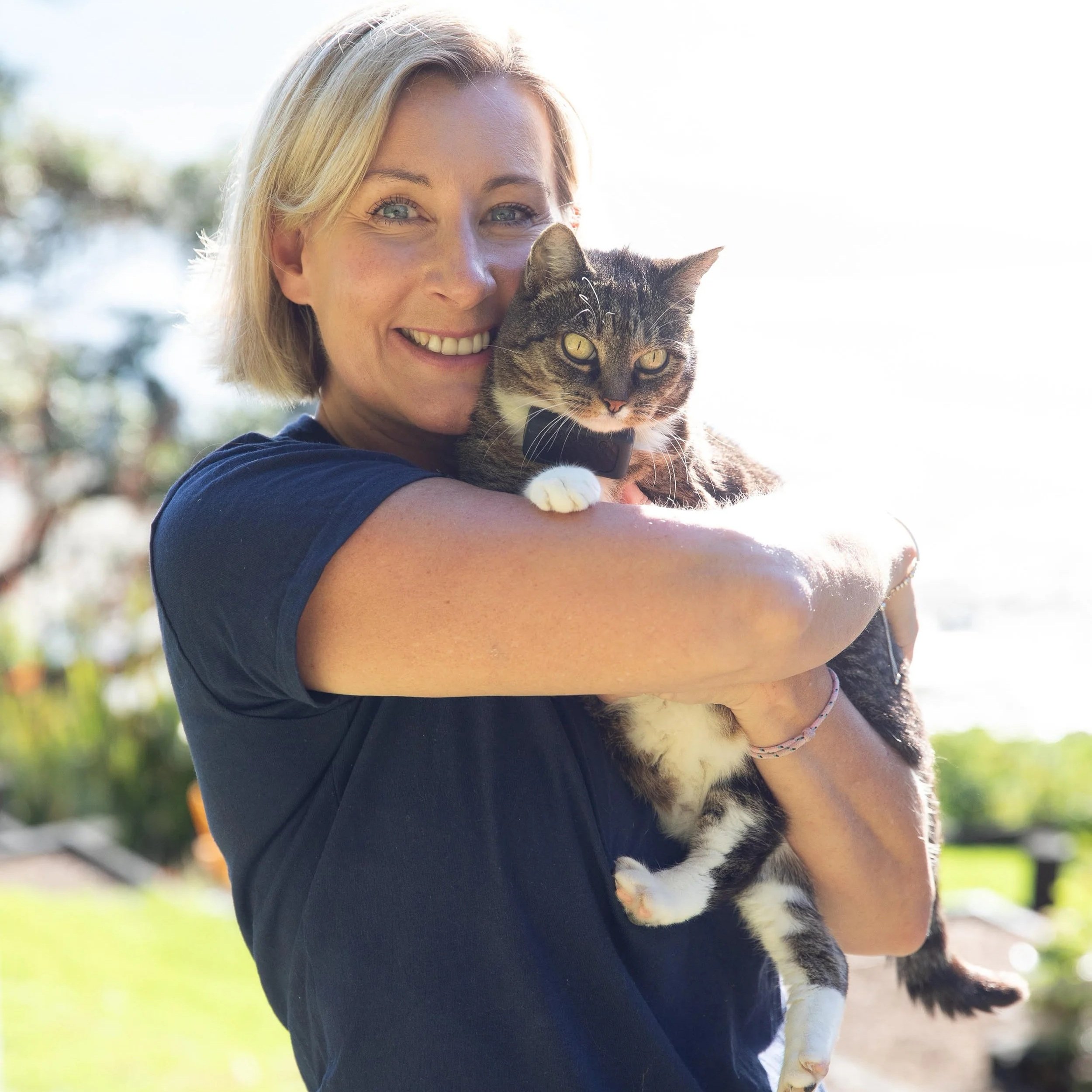 A woman with blonde hair smiling and holding a gray and white tabby cat outdoors on a sunny day.