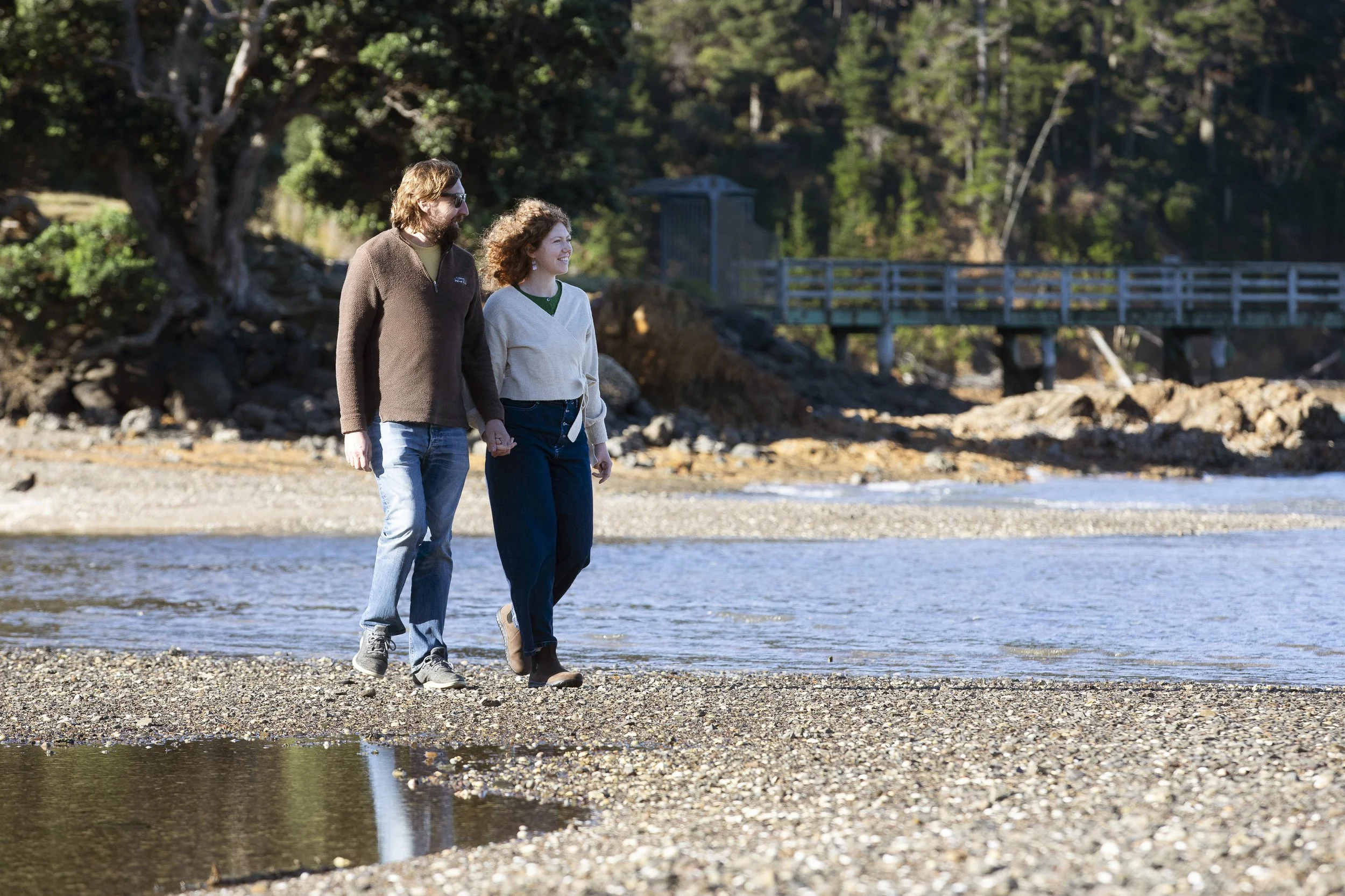 A man and woman walking hand in hand along a riverbank, with a forest and a small bridge in the background.