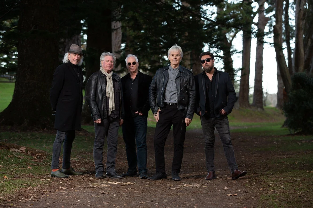 Five men from the band Space Waltz standing outdoors on a dirt path among tall trees, dressed in dark casual clothing with some wearing sunglasses and leather jackets.