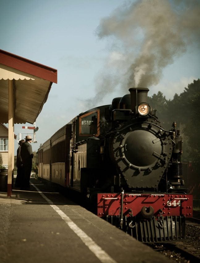 A black steam locomotive train pulling passenger cars at a vintage train station, with a man waiting at the platform and smoke billowing from the chimney.