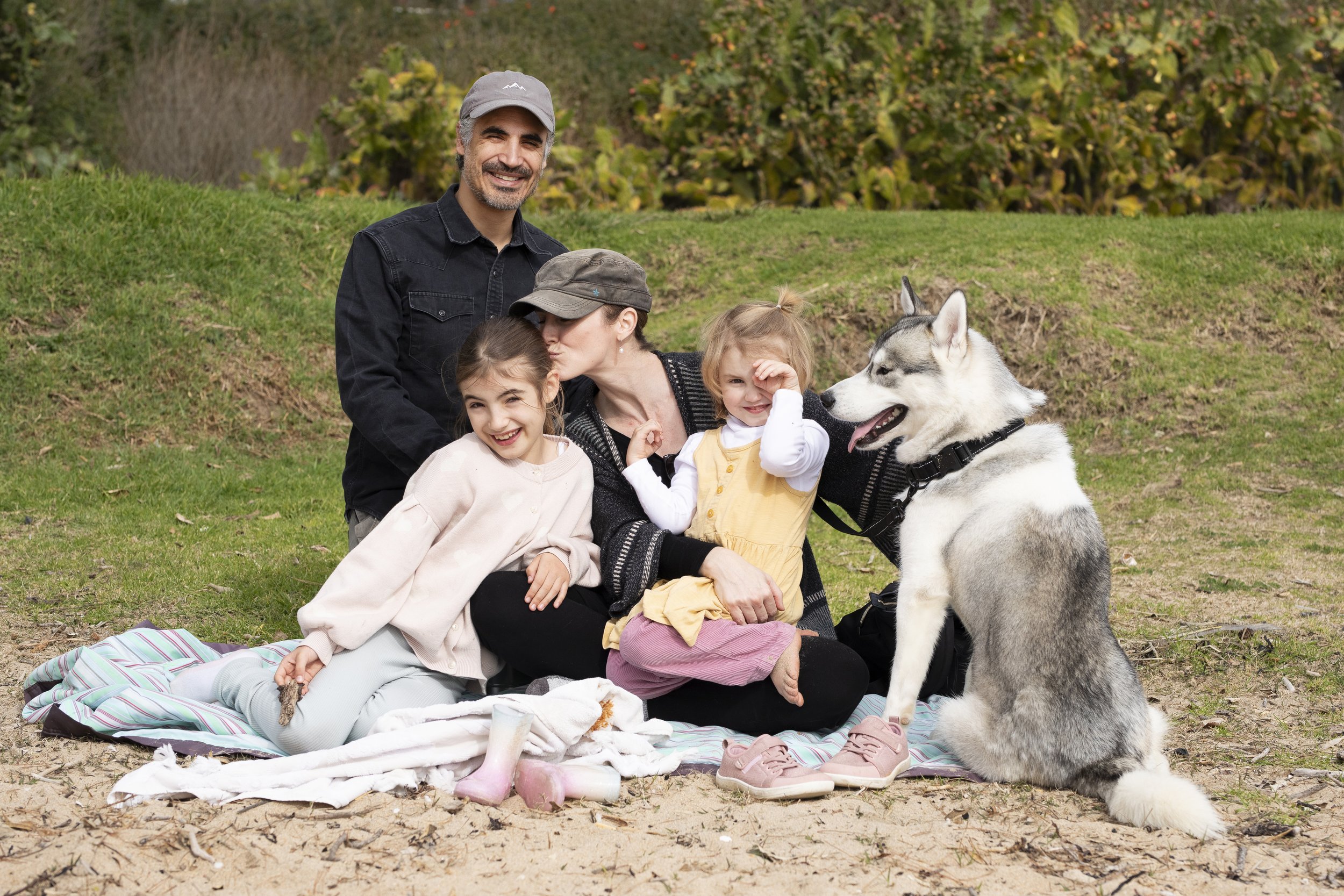 A young family including 2 children, 2 adults and a Siberian husky dog sitting on blankets on the grass at Oneroa Beach on Waiheke Island.