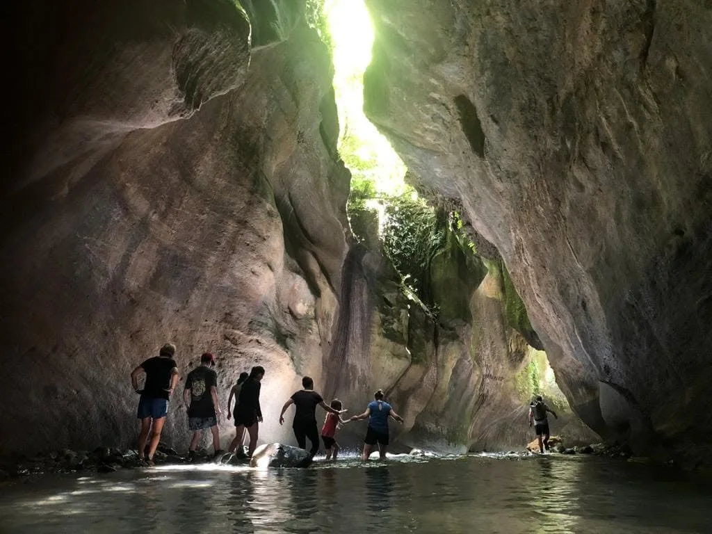 People hiking and holding hands in a river inside a large canyon or cave with high walls and sunlight coming from above.