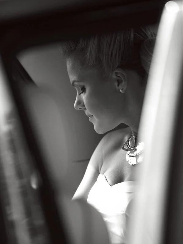 Black and white photograph of a smiling bride at a wedding on Waiheke Island with her eyes closed seen through blurred objects in the foreground.