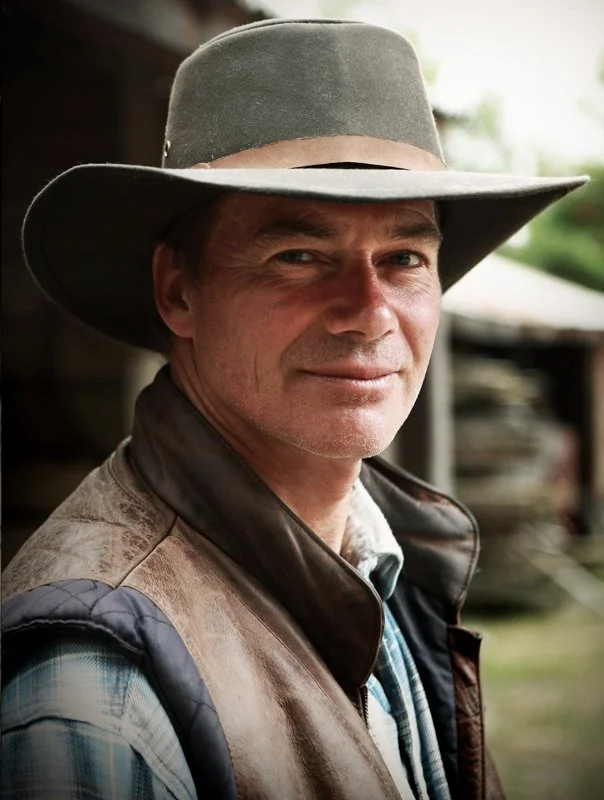 A man wearing a cowboy hat, a leather vest and a plaid shirt, smiling outdoors.