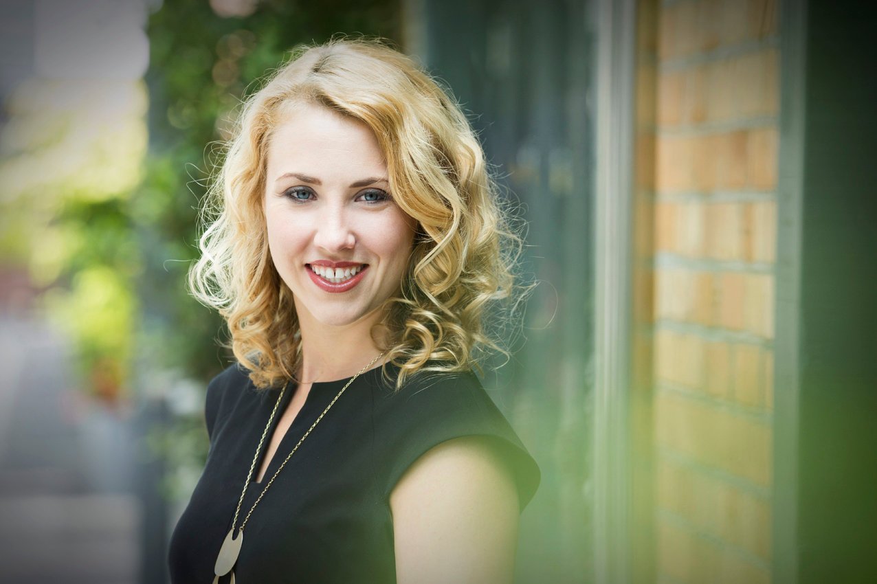 A woman with blonde curly hair smiling outdoors, wearing a black top and a gold necklace, with a brick wall and trees in the background.