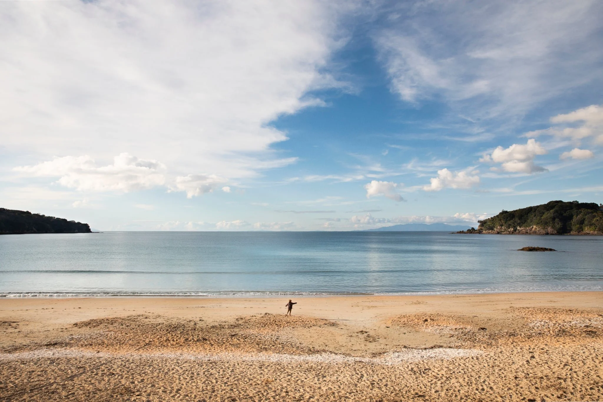 A small child standing by the sea at Oneroa Beach on Waiheke Island. The sky is blue with light white clouds and the ocean is calm.