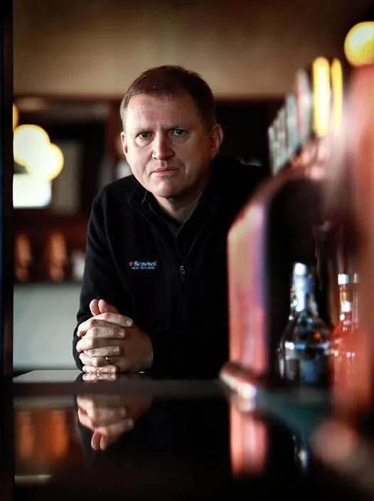 A man with short light brown hair and blue eyes looking directly at camera, seated at a bar or counter, with blurred bottles and lights in the background, wearing a black zip-up jacket.