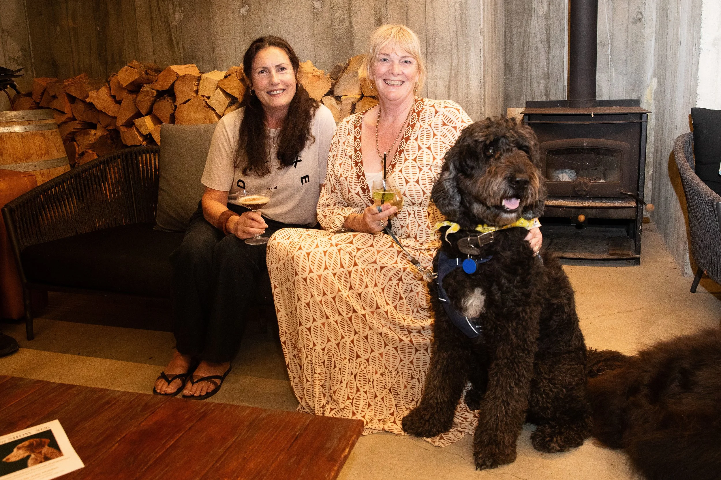 Two women sitting on a black couch, each holding a drink, with a large black dog in front of them. Behind them are stacked firewood and a wood stove, in a rustic setting.