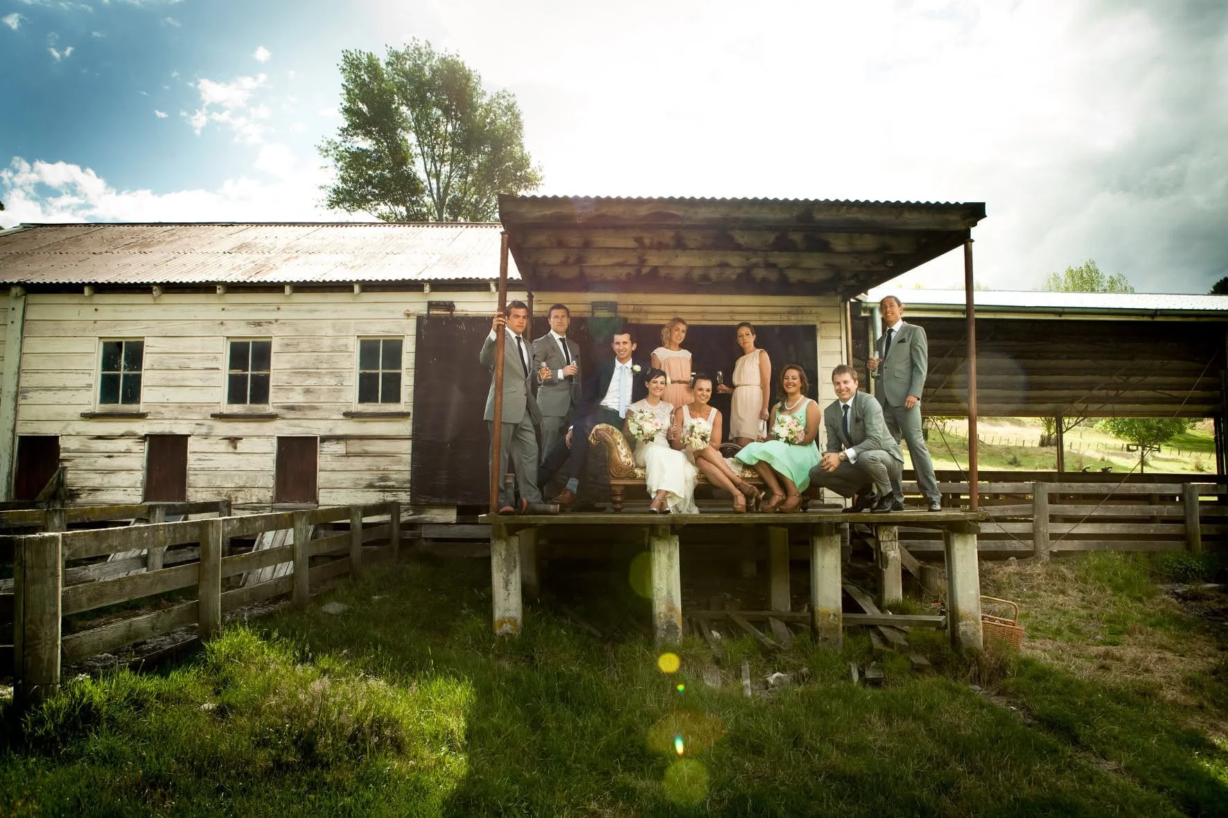 A group of people in formal attire on a rustic wooden porch with a barn in the background, celebrating a wedding outdoors.