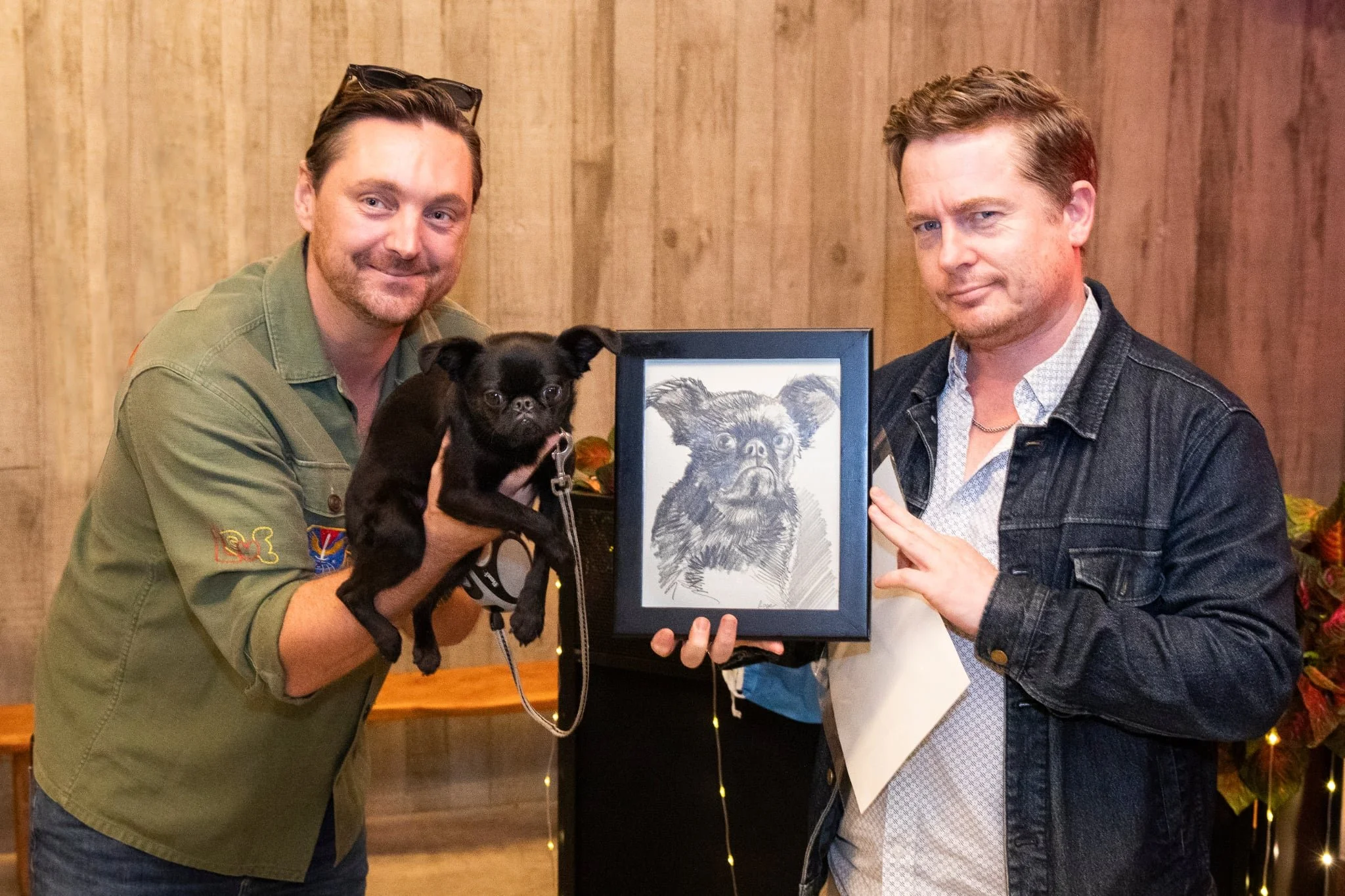 Two men at the Wild Estate on Waiheke Island. One is holding a framed drawing of a dog and one holds the small black dog from the drawing. 