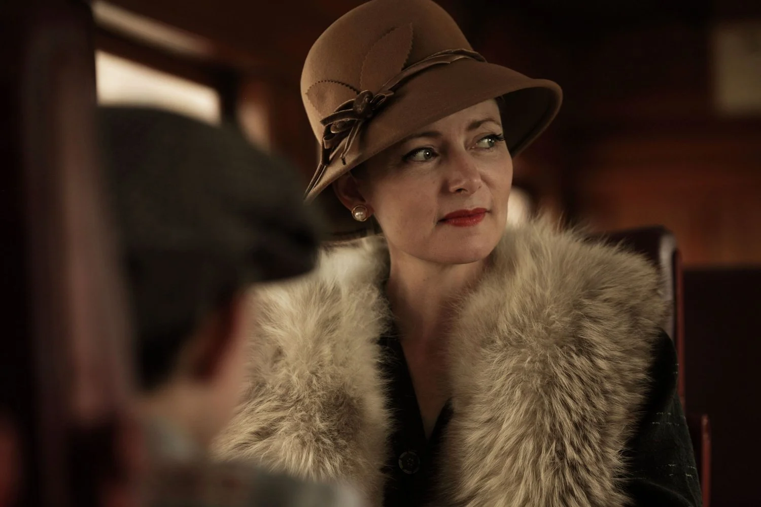 A woman wearing a tan vintage hat with a ribbon and feather, a pearl earring, a fur stole, and a black dress, sitting in a wooden chair in a dimly lit room with a warm, nostalgic atmosphere.