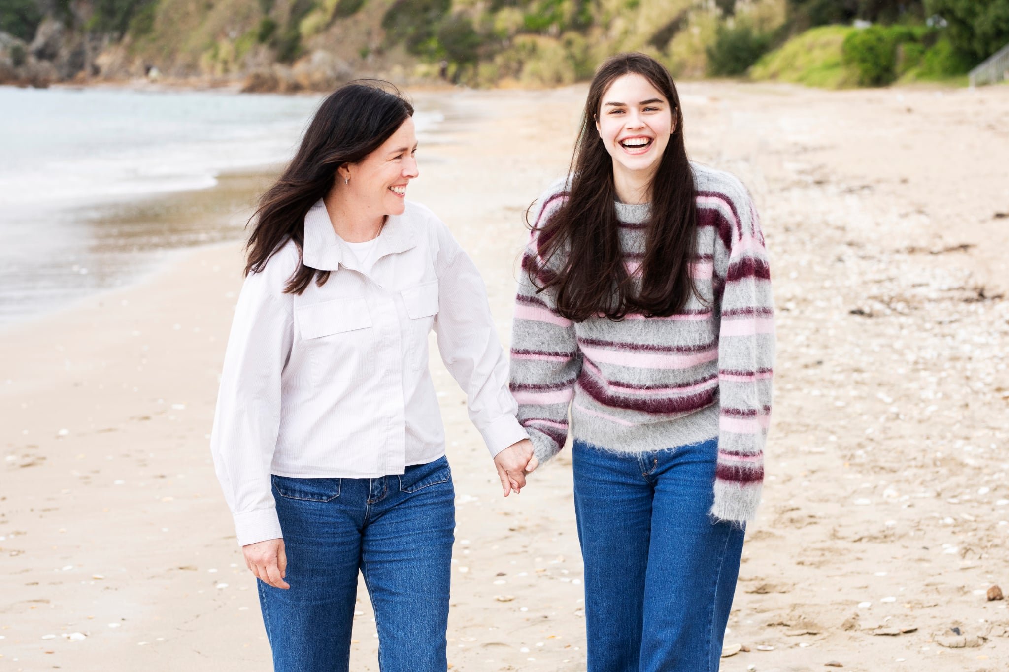 Mother and daughter holding hands and walking on a Oneroa Beach on Waiheke Island with rocks and greenery in the background.
