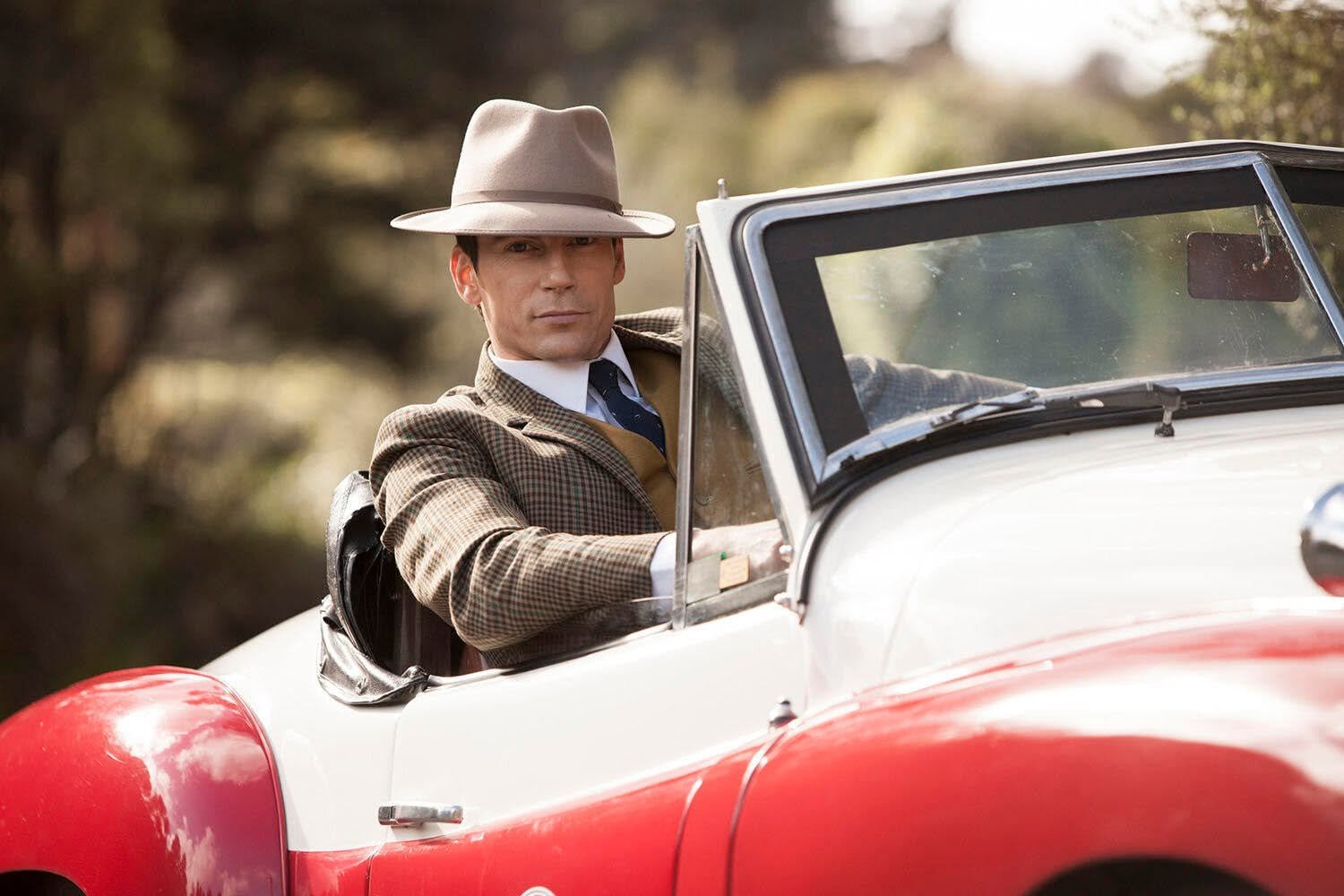 A man in vintage attire, including a tan fedora hat and brown checkered blazer, sits in a vintage red and white convertible car outdoors with a blurred natural background.