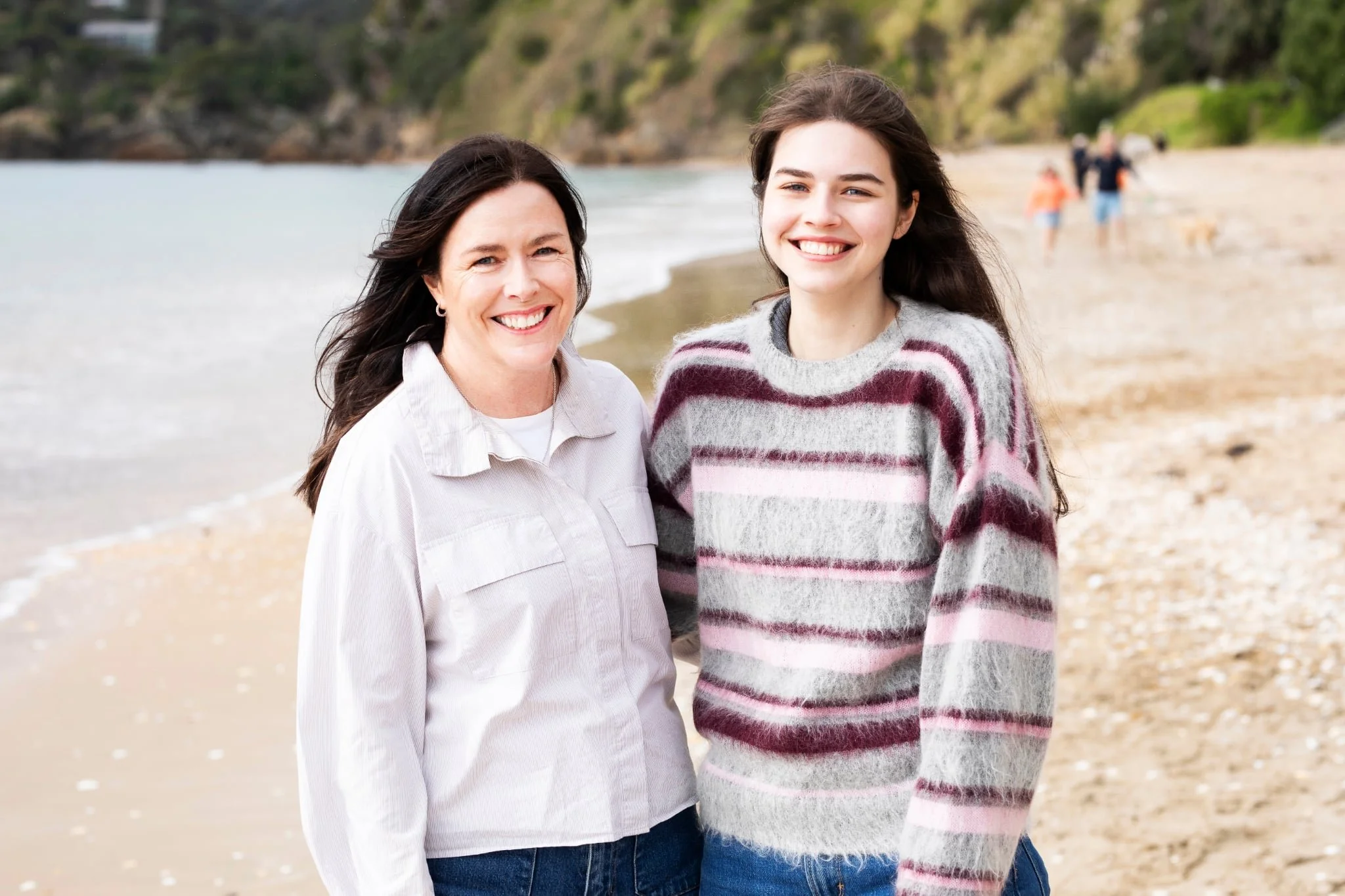 Mother and daughter on Oneroa Beach on Waiheke Island with sand and ocean, cliffs and trees in the background.
