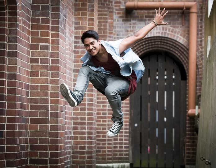 A young man jumping in a narrow alleyway between brick buildings, smiling at the camera.